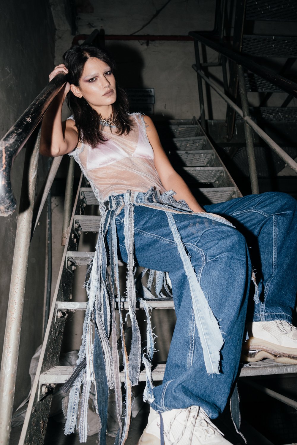 A woman with dark hair and makeup sitting on a metal staircase in an industrial setting, wearing a sheer top and baggy jeans with fabric strips attached.