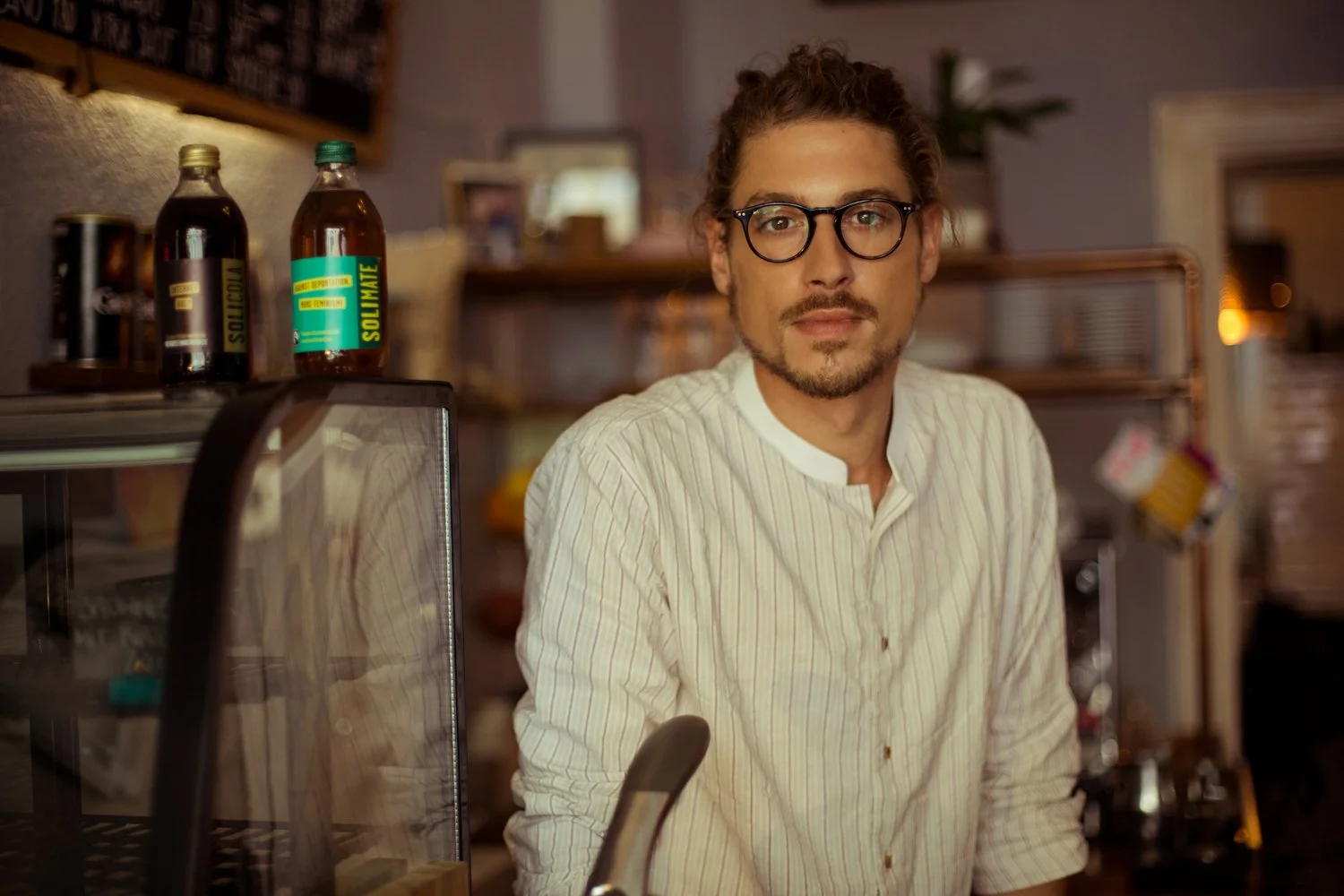 A man with glasses and a beard, wearing a white striped shirt, stands behind a counter in a cafe or restaurant.