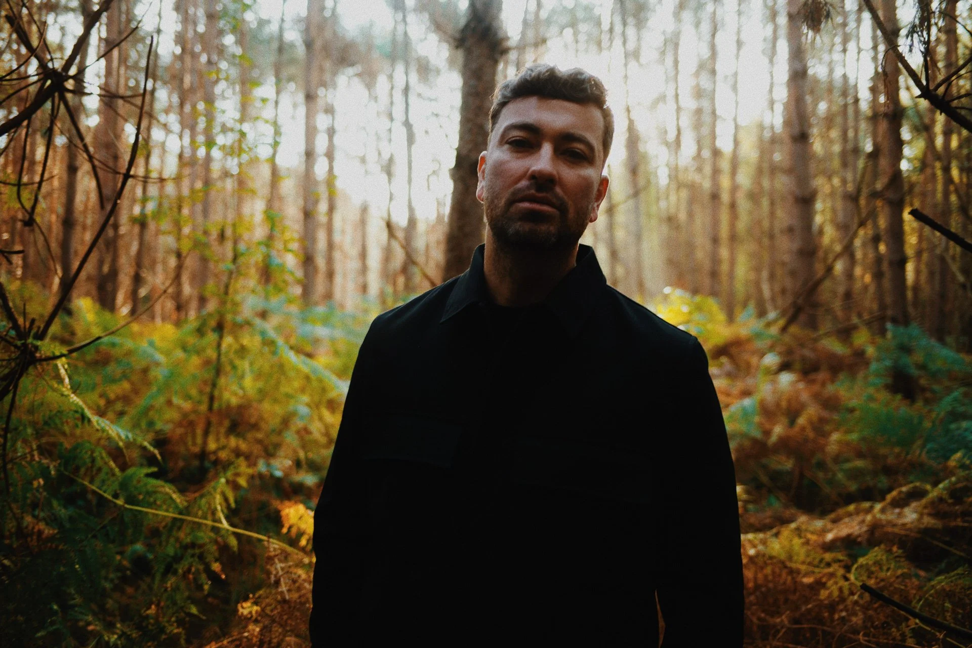 A man with dark hair and a beard stands in a forest with trees and ferns, wearing a black shirt.