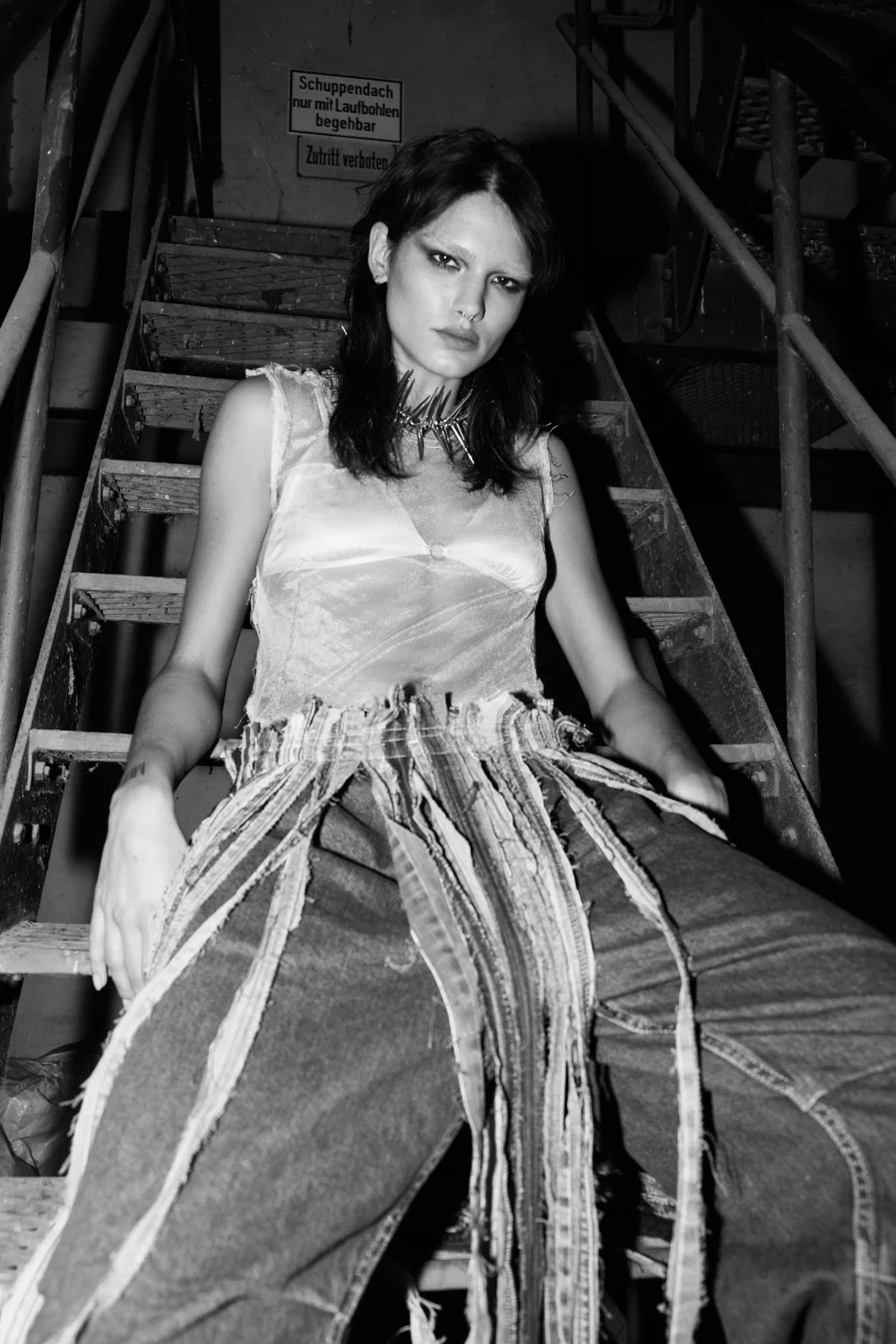 Black and white photo of a young woman sitting on stairs with a serious expression. She has dark hair, wears a spiked necklace, a sleeveless top, and baggy pants with multiple strips of fabric hanging down.