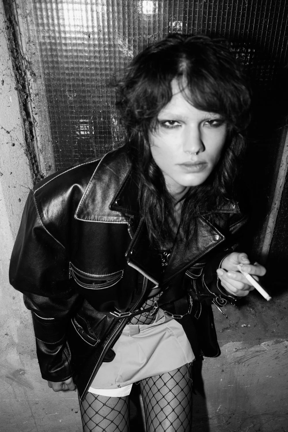 Black and white photo of a woman with intense expression, wearing a leather jacket, fishnet stockings, and holding a cigarette, standing against a textured wall.