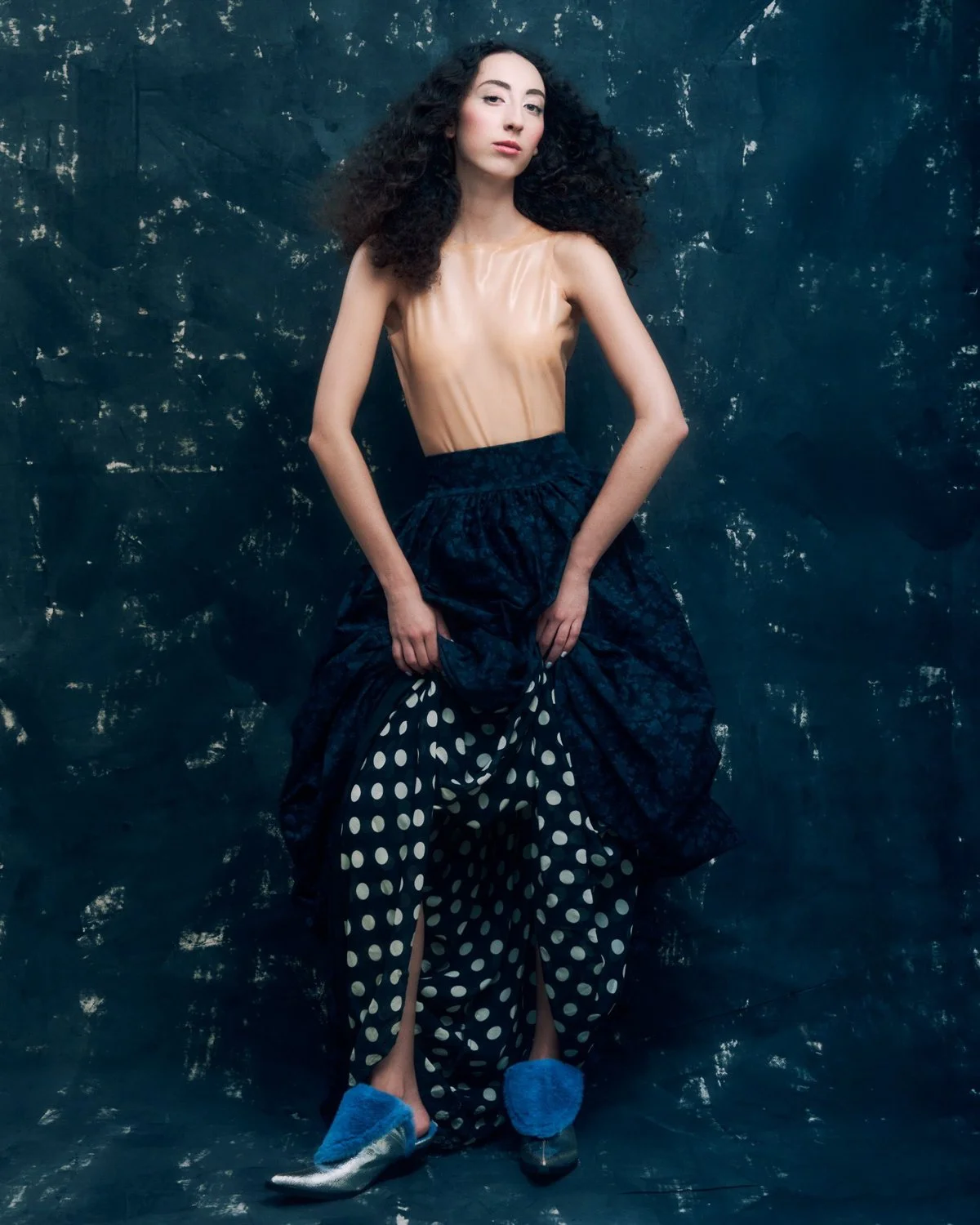 A young woman with long curly black hair, wearing a sheer tan top, a black skirt with polka dots, blue fuzzy slippers, and holding up her skirt against a dark textured background.