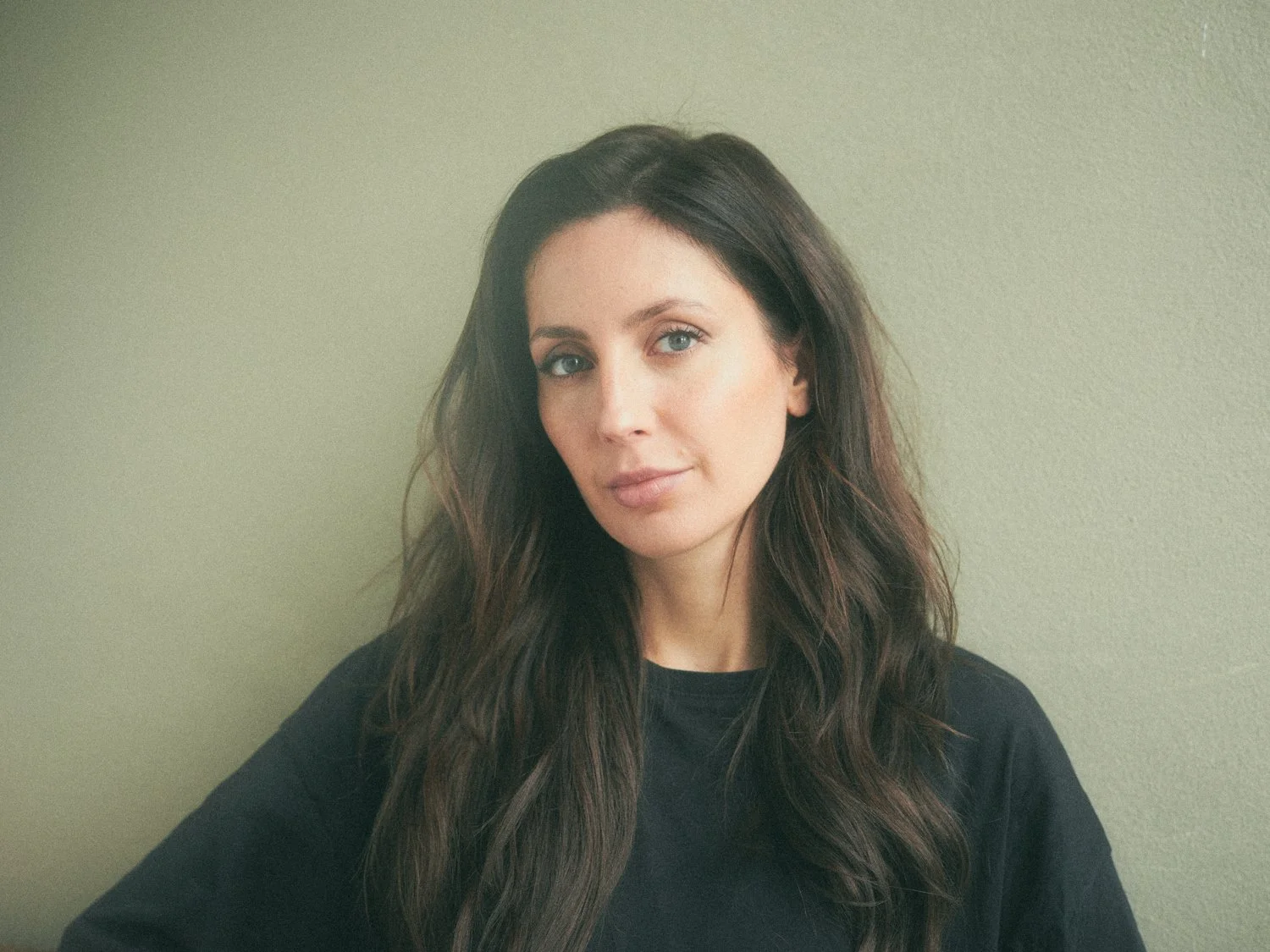A woman with long brown hair and blue eyes looking at the camera against a light green wall.