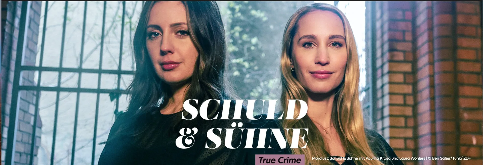 Two women posed side by side with a fence and trees in the background, promoting a show titled 'Schuld & Sühne'.
