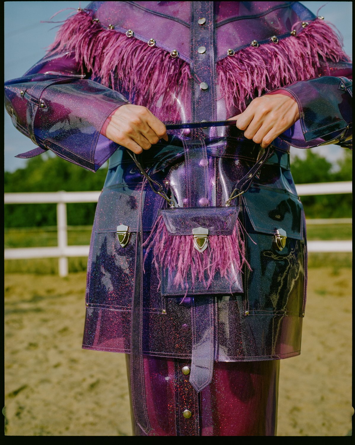 Close-up of a person wearing a shiny, galaxy-themed outfit with pink feather details, holding a matching transparent bag in front of a rural background.