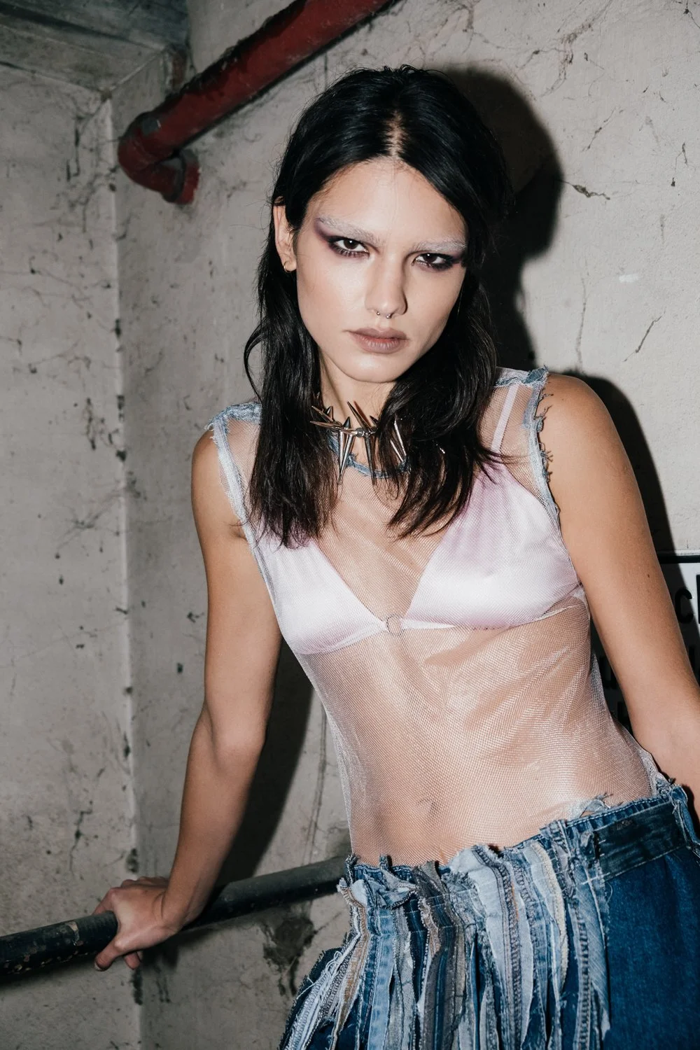 A young woman with black hair, makeup, and piercings, wearing a sheer pink top and ripped jeans, standing against a concrete wall with a red pipe.