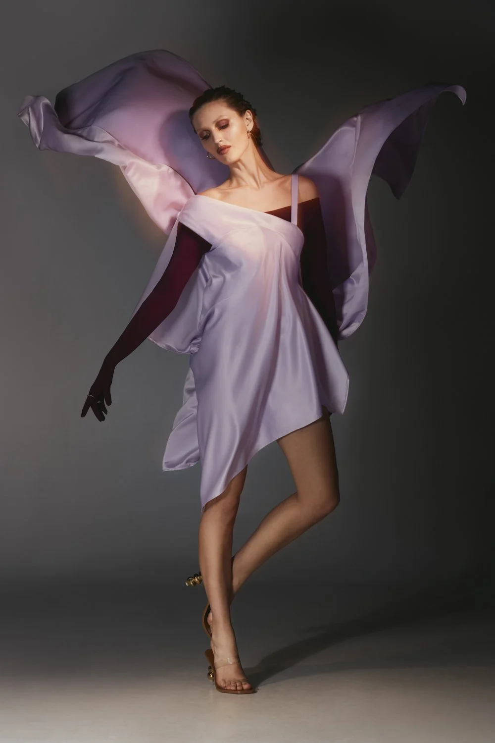 A woman in a flowing lavender and black dress, striking a graceful pose against a dark background, with heels and makeup.
