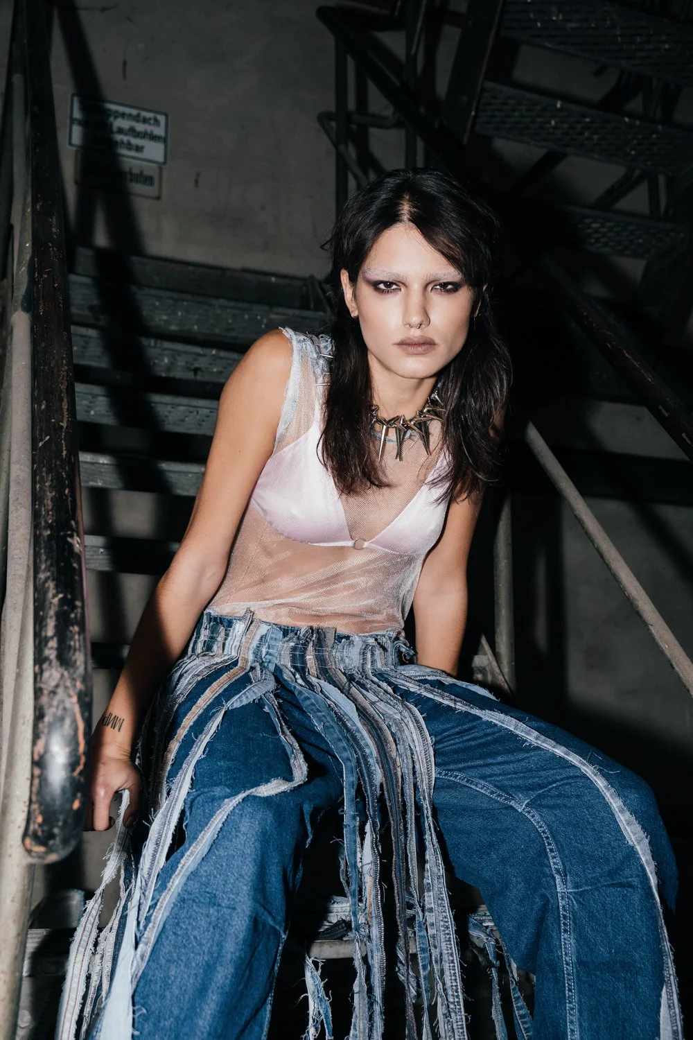 A young woman with dark hair and makeup sitting on a metal staircase, wearing a sheer pink top, ripped jeans with hanging strips of denim, and a spiked necklace.