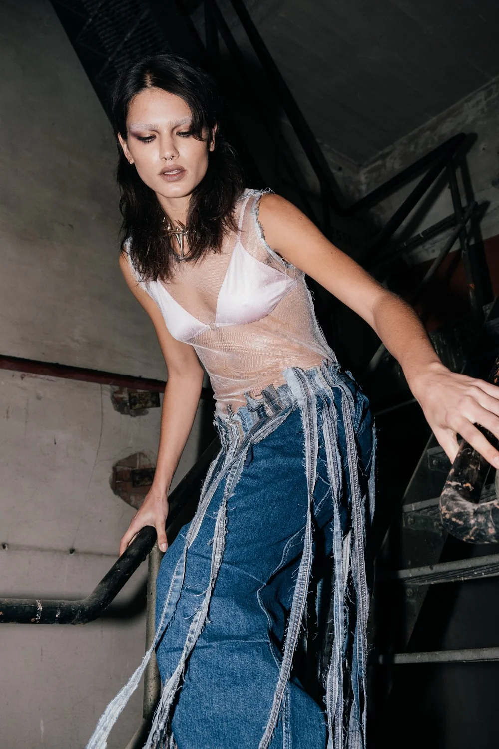 A young woman with dark hair and makeup, wearing a sheer tank top and patched blue jeans, leaning on a railing in an industrial setting.