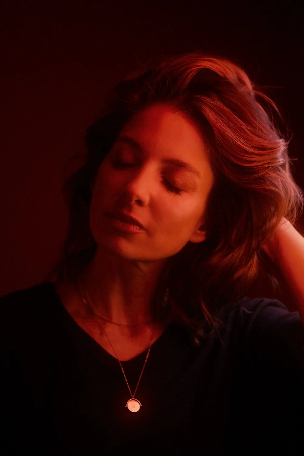 A woman with wavy brown hair and closed eyes, illuminated by red lighting, wearing a black top and a gold necklace.
