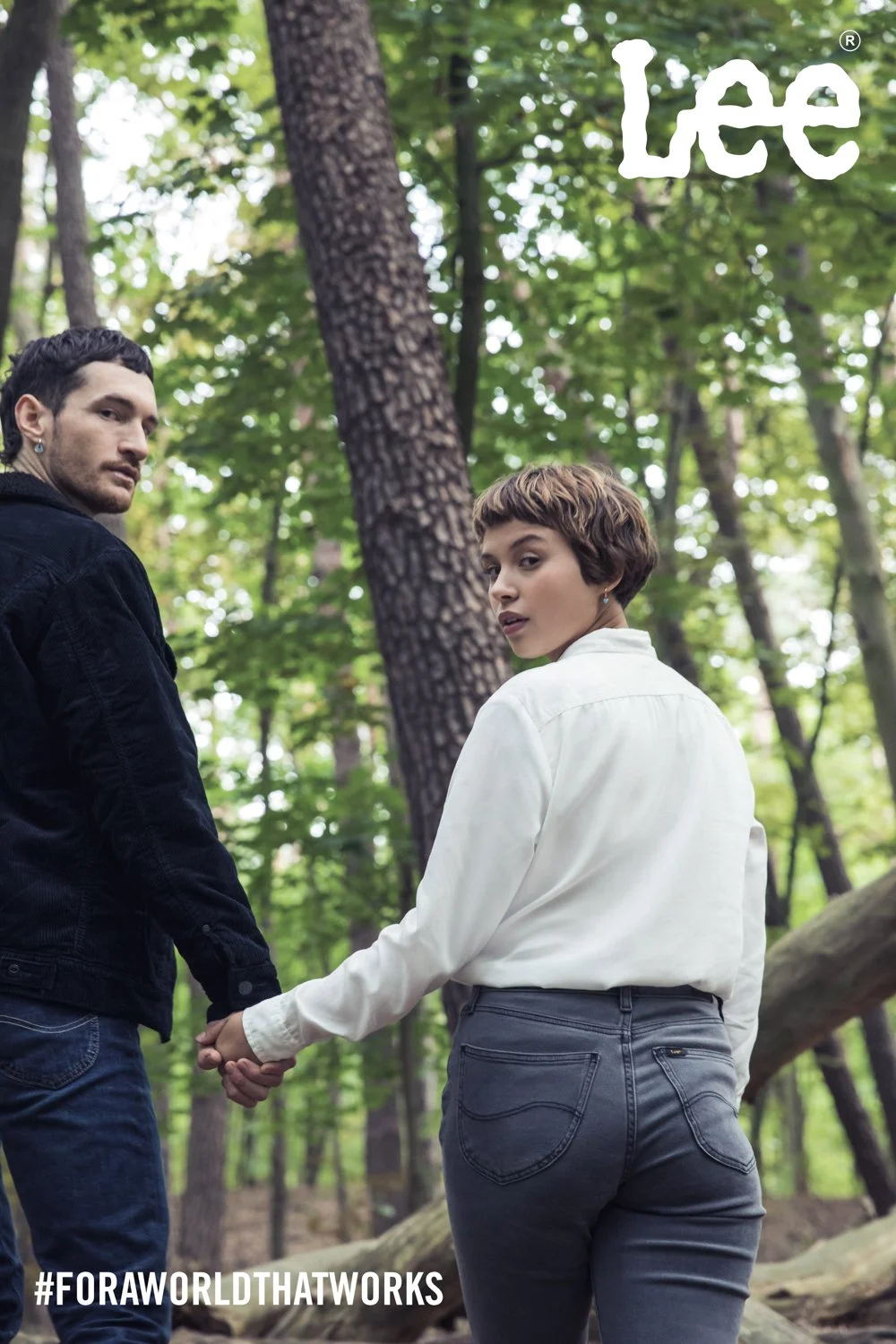 A man and a woman holding hands in a forest, looking back over their shoulders. The woman has short brown hair and is wearing a white shirt and gray jeans, while the man has dark hair and is wearing a black jacket and jeans. The background consists o