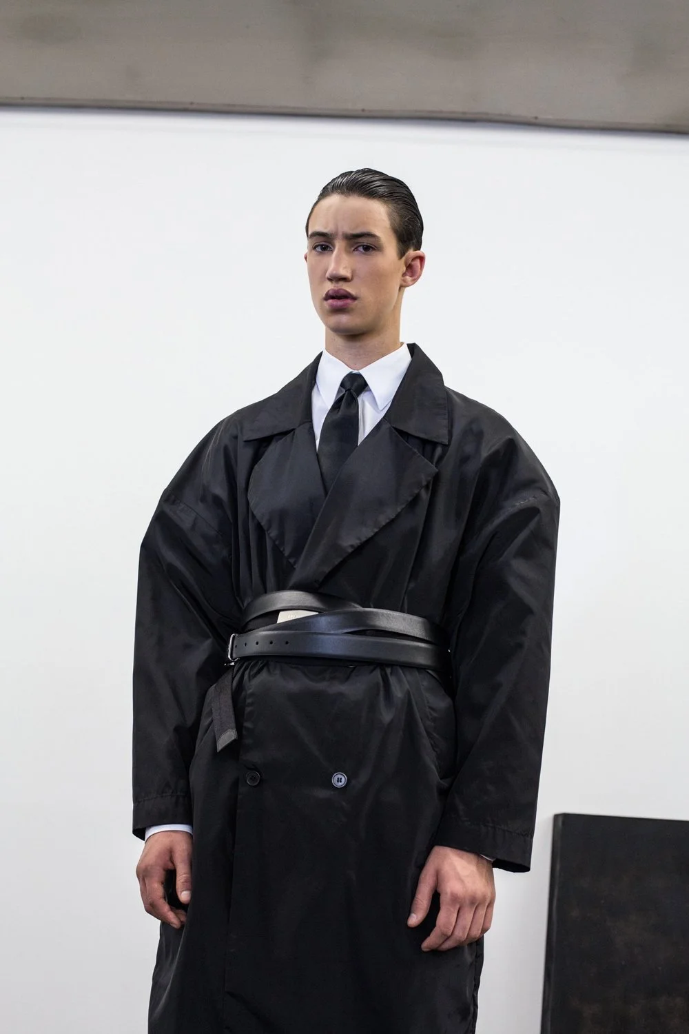 Young man in formal black attire with coat and tie, standing against a plain white background, looking at the camera with a neutral expression.