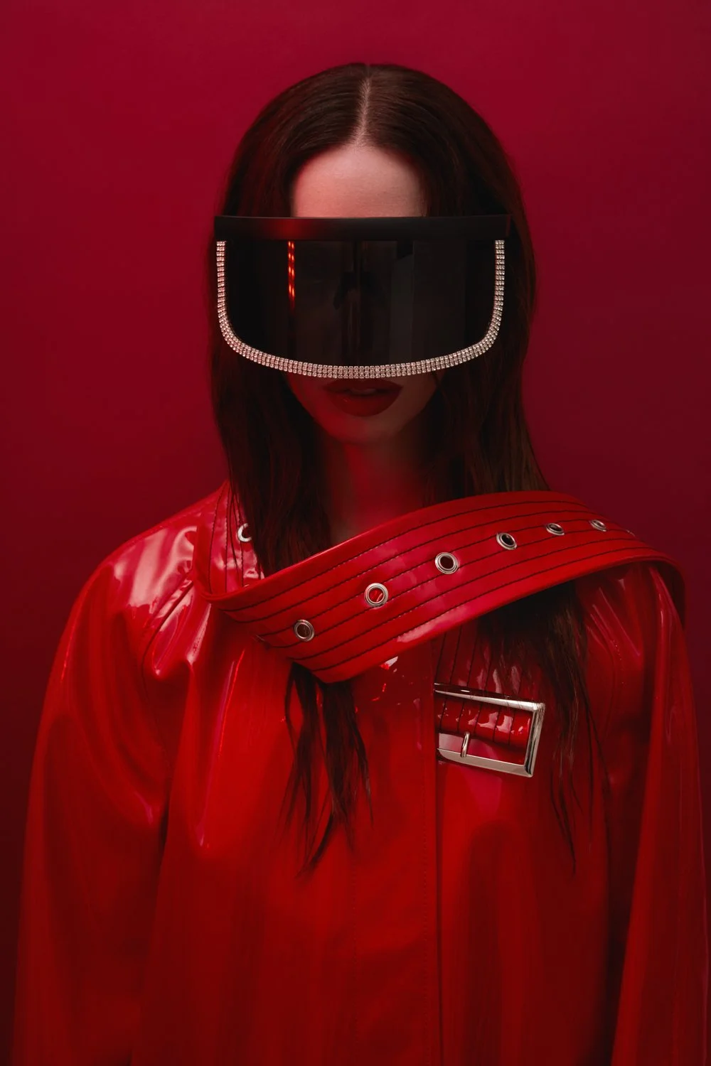 A woman with long dark hair wearing a red vinyl jacket, large black visor sunglasses with rhinestones, and a red background.