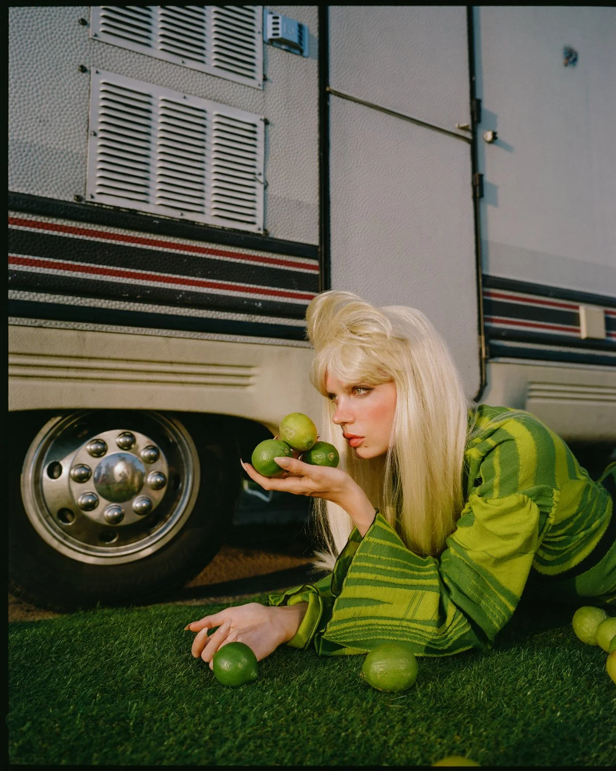 A woman with long blonde hair and a green striped jacket is lying on grass, holding a bunch of limes near her face, with some limes scattered on the grass near her. She is positioned next to a large vehicle, possibly a bus or RV.