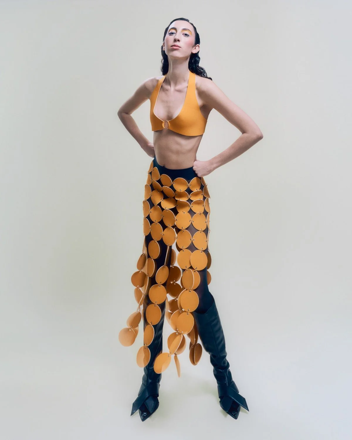 Person wearing a distinctive fashion outfit consisting of a mustard yellow bra top and black pants with a fringe made of large circular orange discs, paired with black pointed boots, standing against a plain light background.