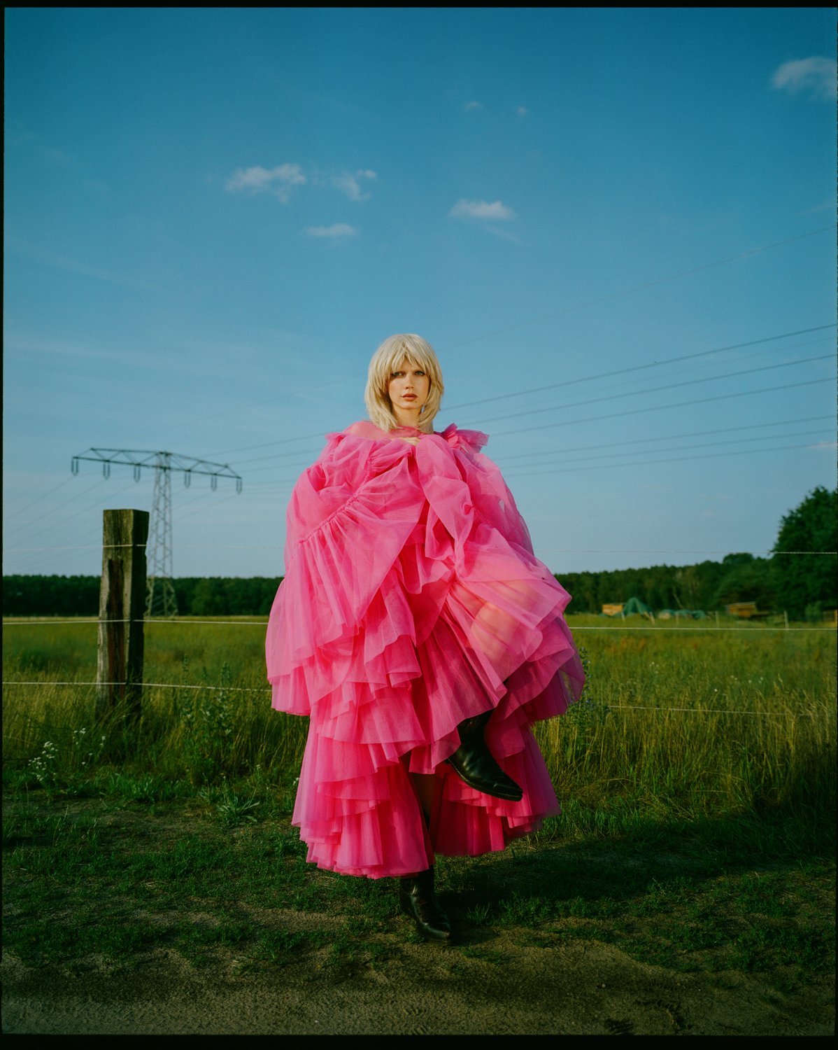 A woman wearing a vibrant pink, layered tulle dress standing outdoors in a grassy field with power lines and green trees in the background under a blue sky.