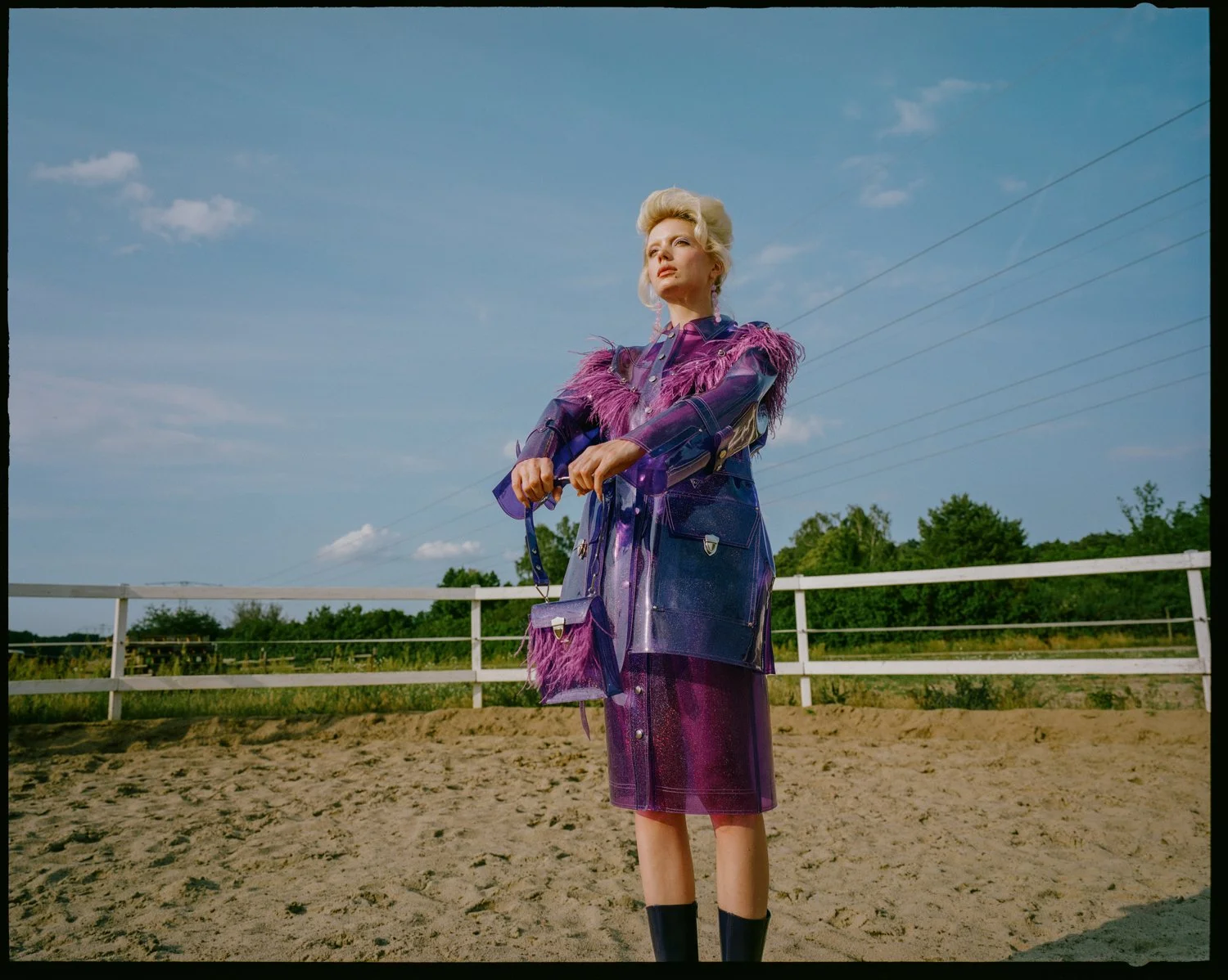 A woman in a purple and pink outfit with feathers, standing on a dirt area with a white fence and green trees in the background, under a partly cloudy sky.