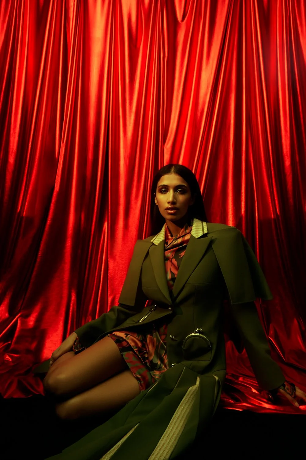 A woman in a military-style blazer with a floral dress and black tights sitting in front of a red satin curtain.