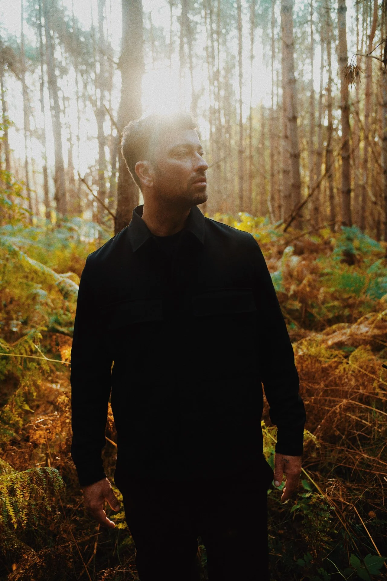 A man in a black jacket standing in a forest with sunlight shining through trees and foliage.