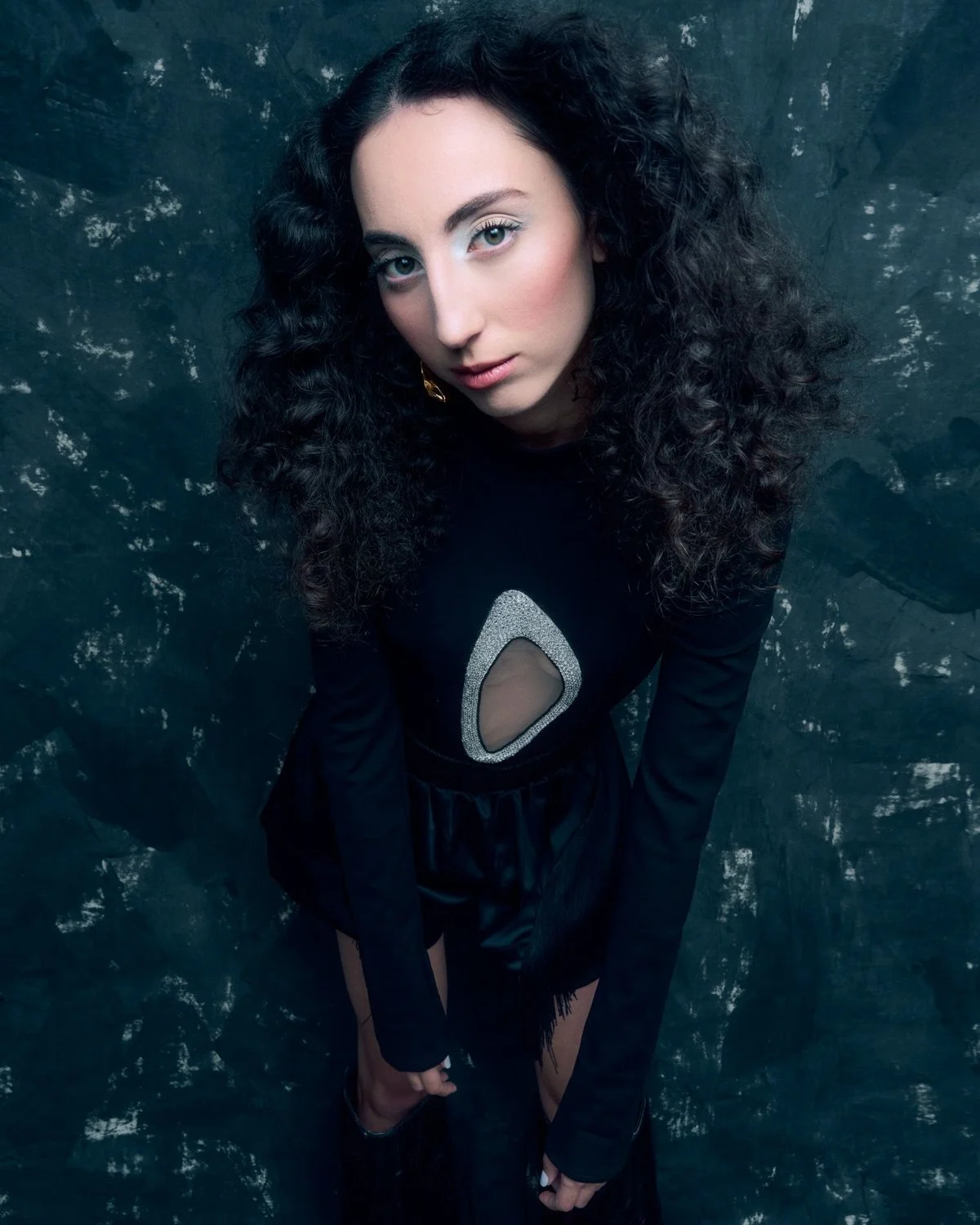 A woman with long curly black hair and light skin looking up at the camera, dressed in a black outfit with a silver and sheer cut-out design on the chest, positioned against a dark textured background.