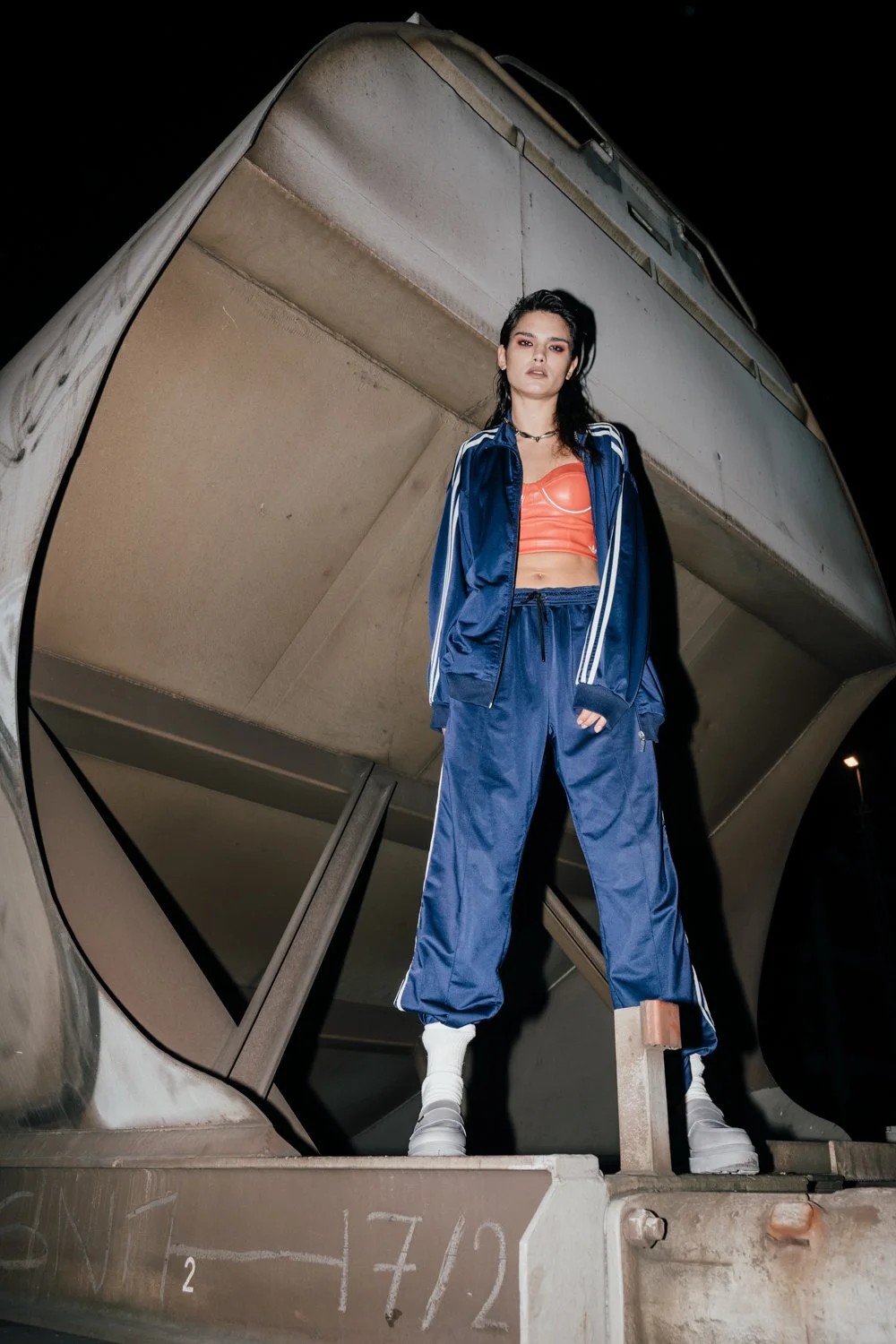 A woman stands on a concrete block in front of a modern, abstract architectural structure at night. She wears a blue tracksuit, a red crop top, white boots, and has short black hair.