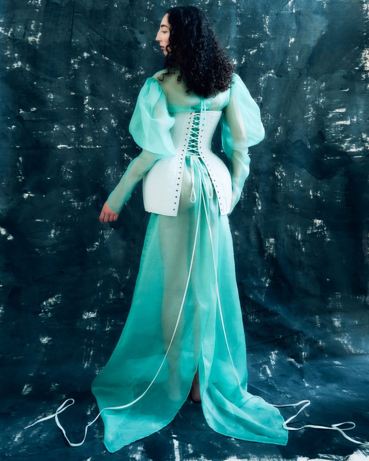 A woman with long curly black hair wearing a sheer turquoise dress with puffed sleeves and a corset back is standing against a dark textured backdrop.