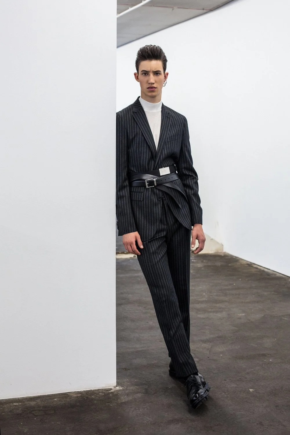 A young man in a black pinstripe suit with a wide belt, white turtleneck, and black shoes, standing in an art gallery or studio space with white walls and concrete flooring.