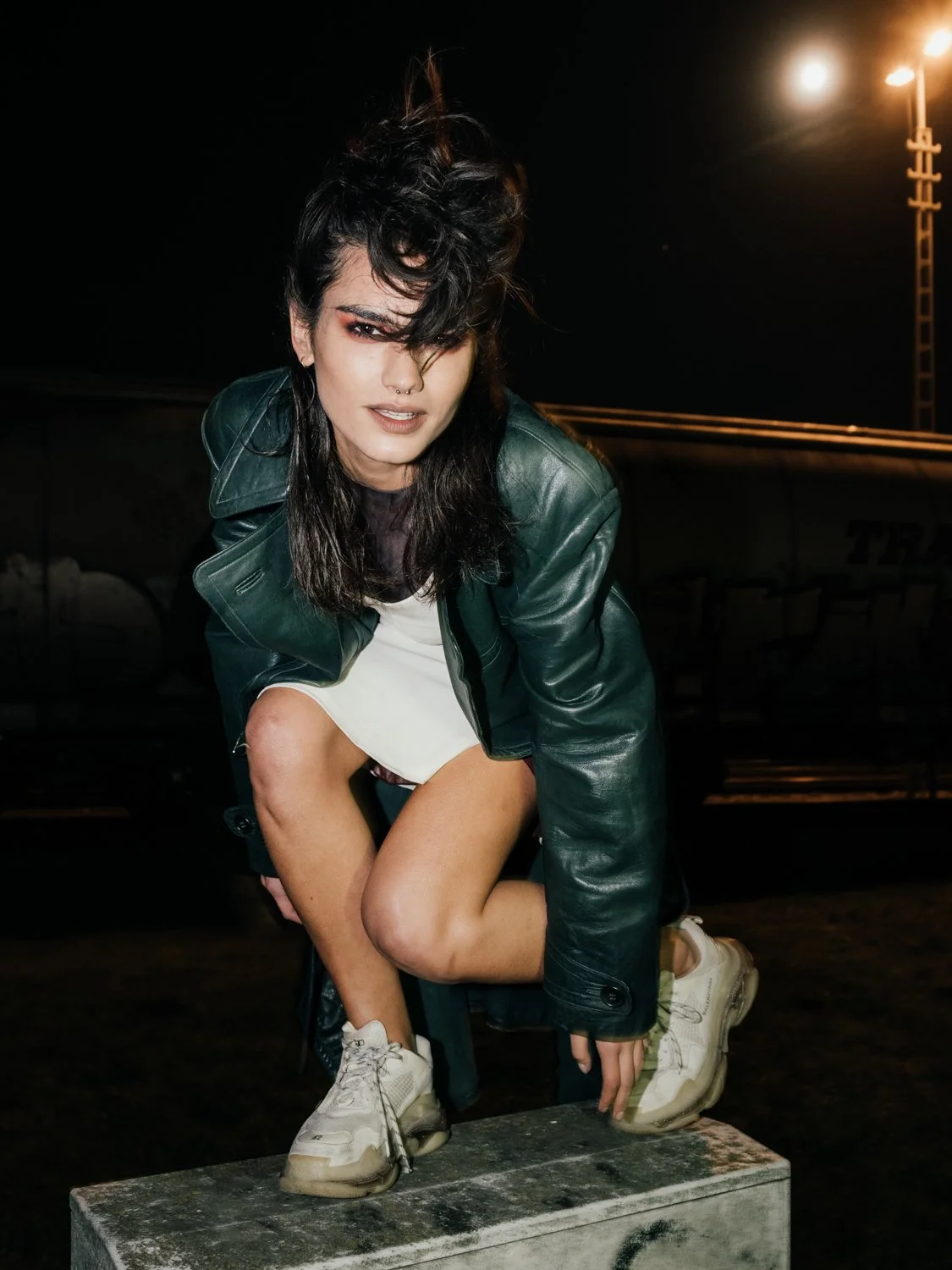 A woman with dark, wavy hair and a septum piercing crouches on a concrete block at night, wearing a leather jacket, white dress, and chunky white sneakers, against a dark background with bright streetlights.
