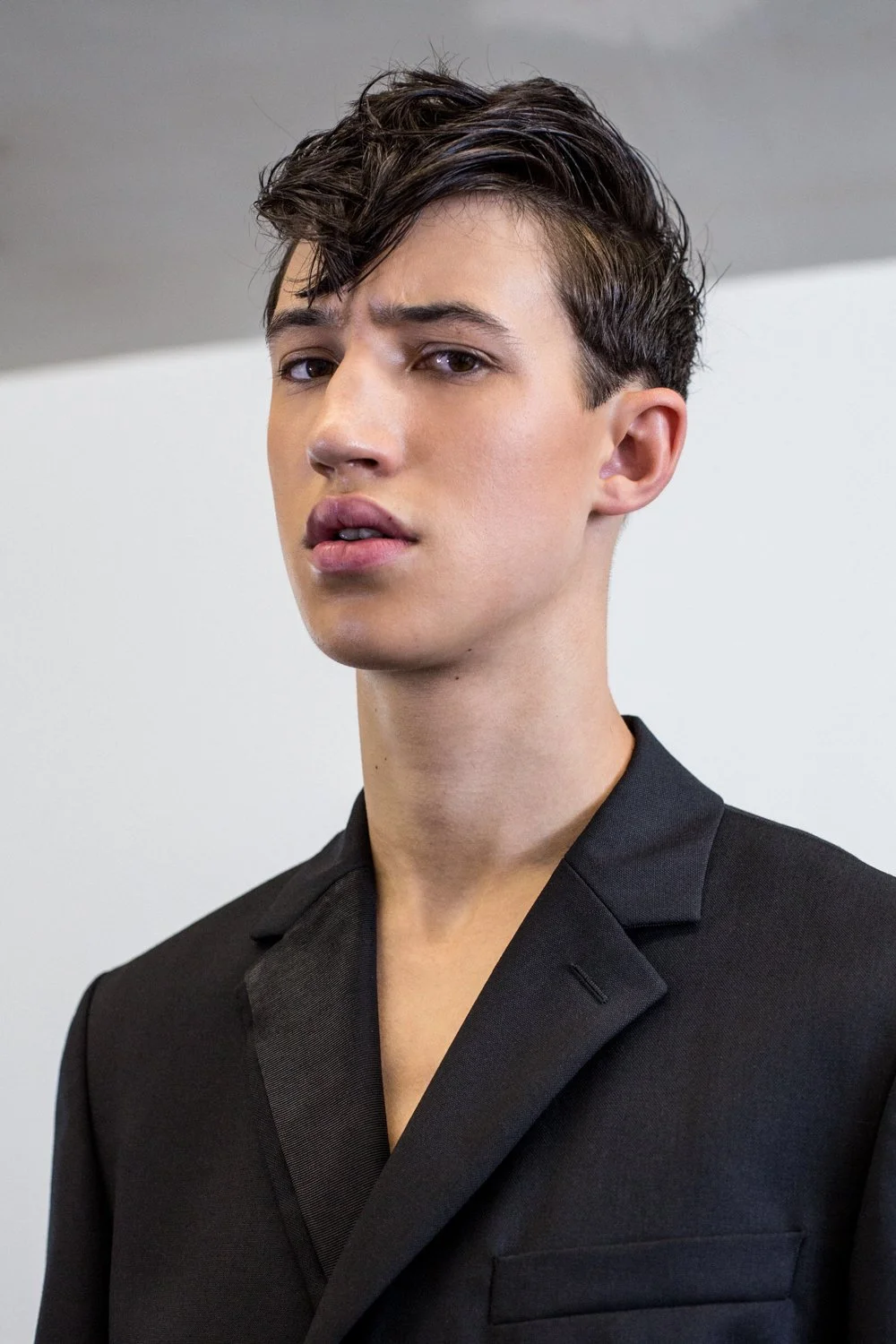 A young man with dark, wavy hair wearing a black blazer, looking slightly to the side with a neutral expression, in front of a plain background.