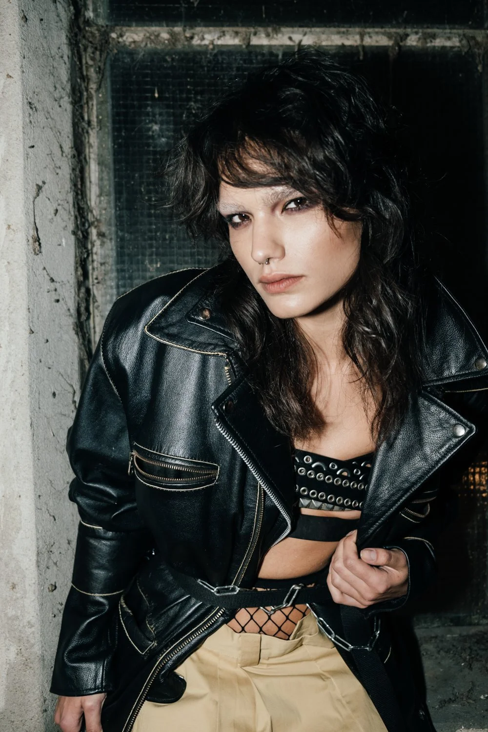 A woman with dark, curly hair wearing a black leather jacket, fishnet top, and beige pants leans against a rough concrete wall in an urban setting.
