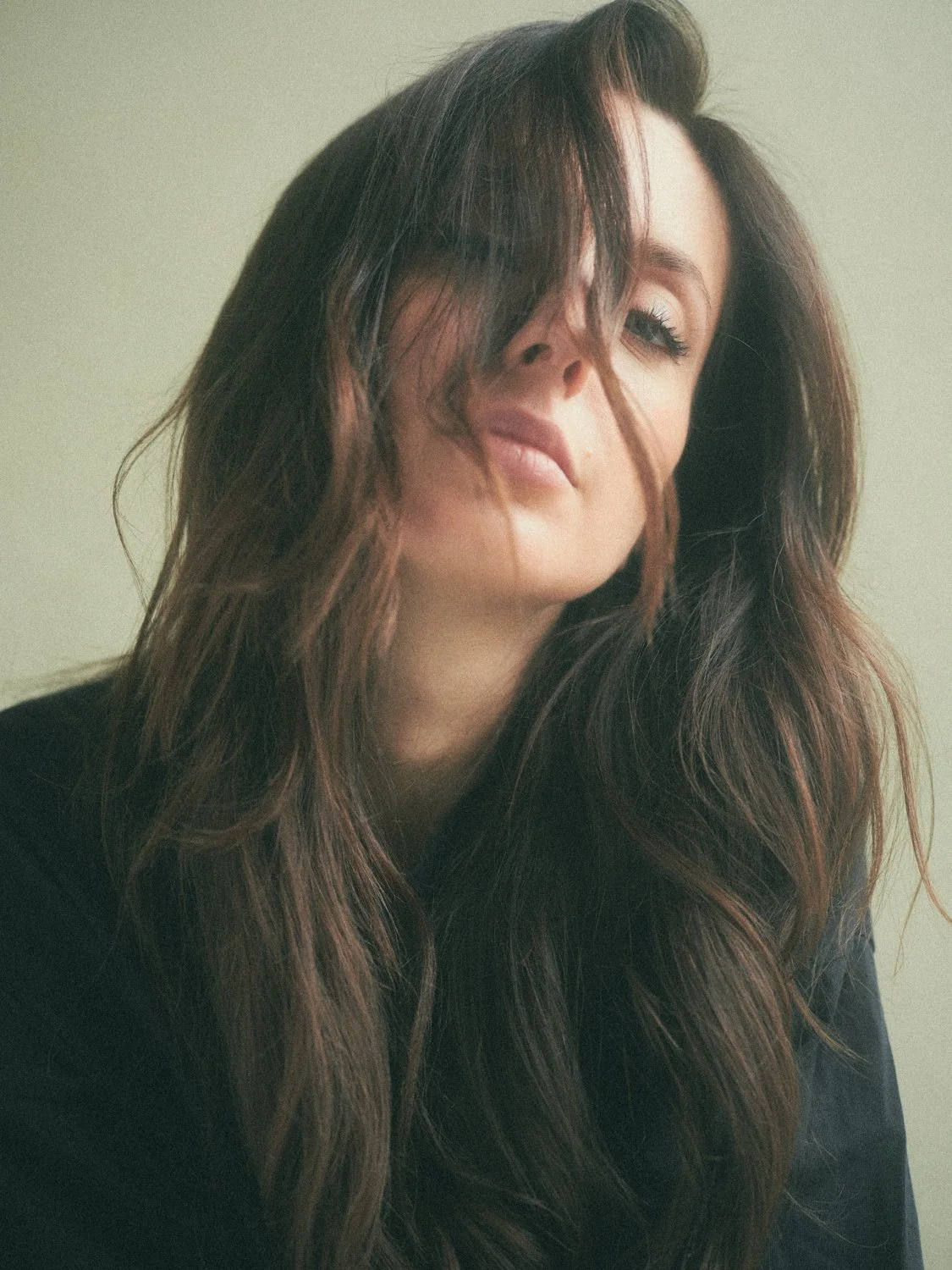 Close-up of a woman with long, wavy brunette hair, partially covering her face, with a neutral background.