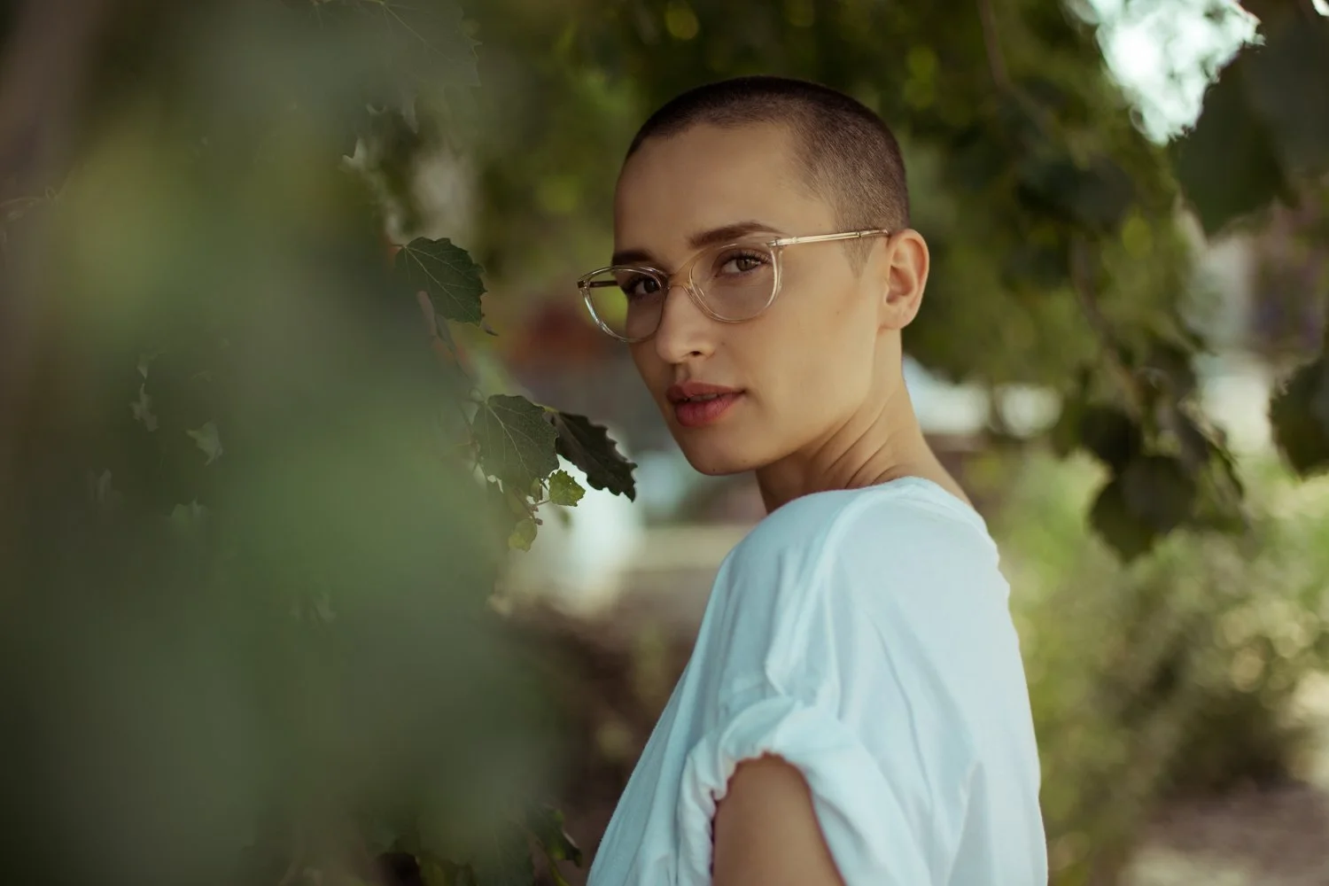 A young woman with short hair and glasses standing outdoors among green leaves, looking at the camera.