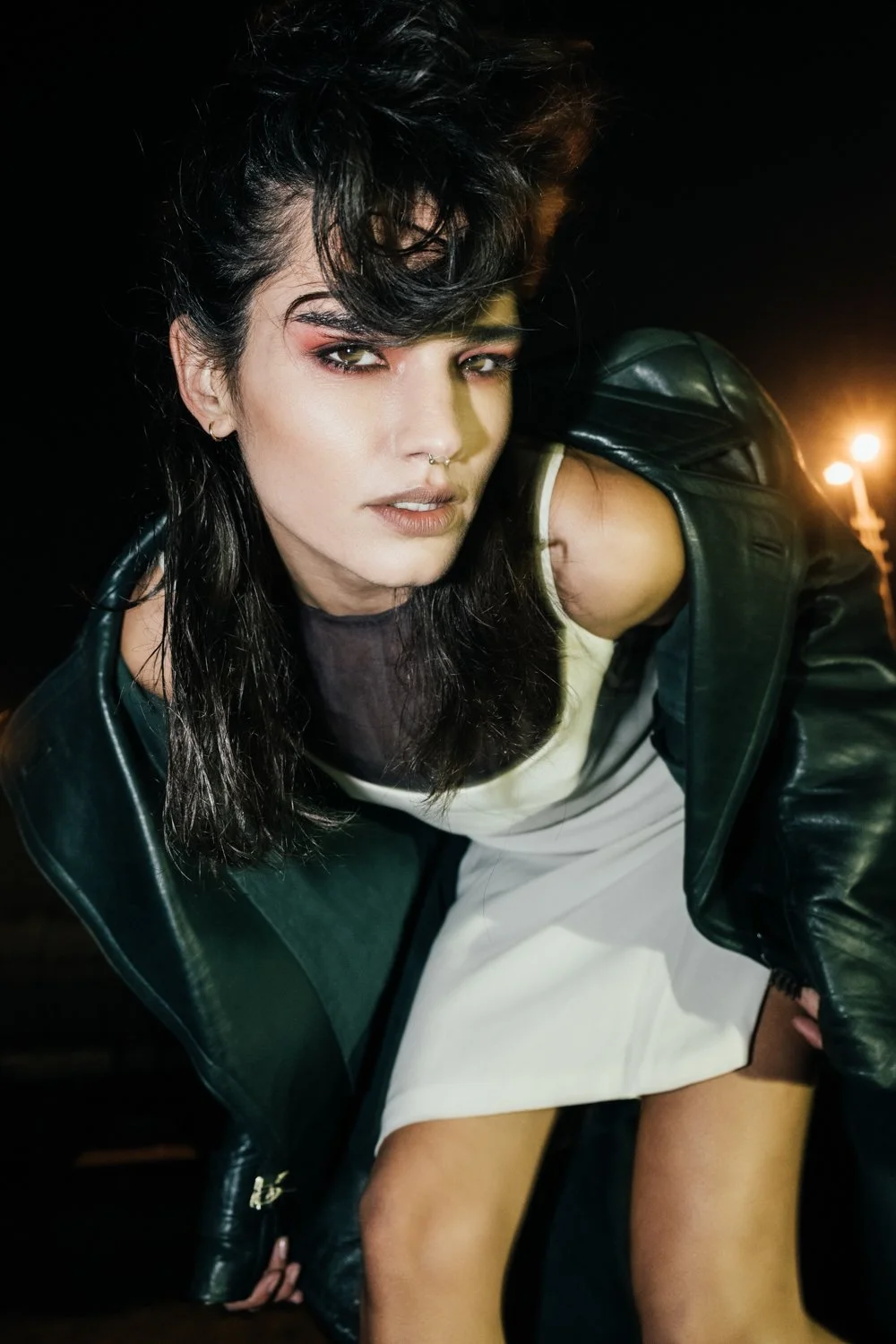 A young woman with dark, styled hair, wearing makeup and a septum piercing, poses outdoors at night in a leather jacket and a white dress with a strap falling off her shoulder.