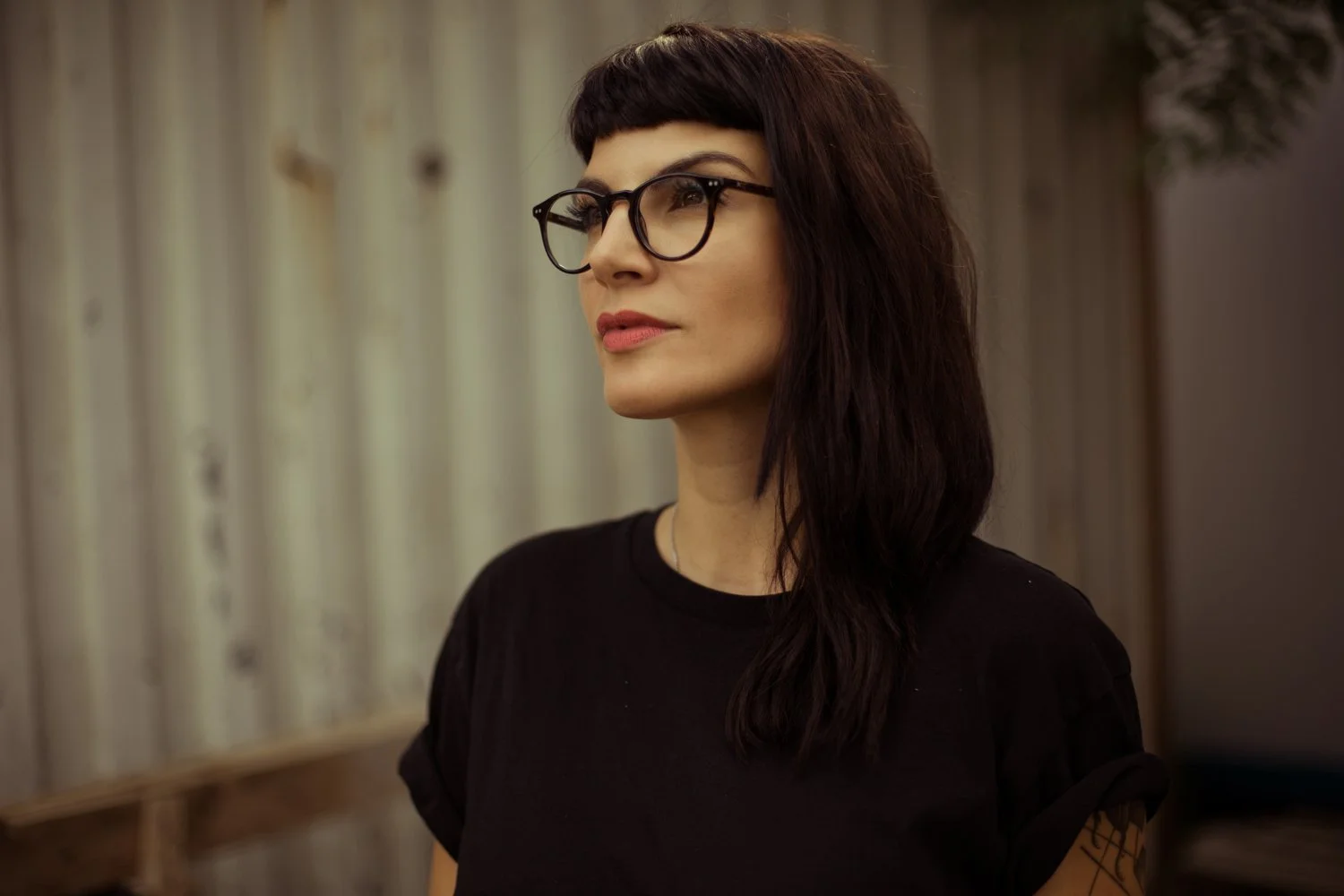 A woman with black hair, glasses, and wearing a black shirt standing by a wooden fence.