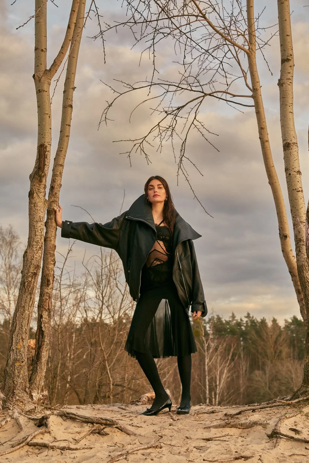 A woman dressed in black, wearing a leather jacket and a pleated skirt, standing among leafless trees outdoors during sunset or sunrise, with a cloudy sky in the background.