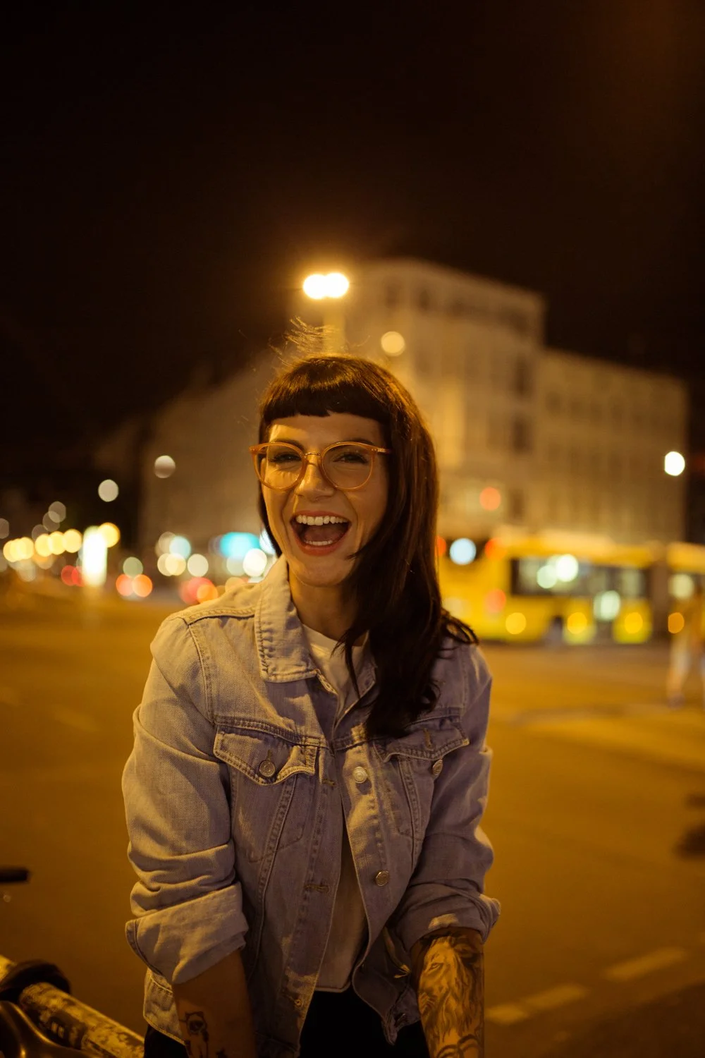 A young woman with dark hair, glasses, and tattoos on her arm, smiling and laughing at night on a city street with blurred lights and a building in the background.
