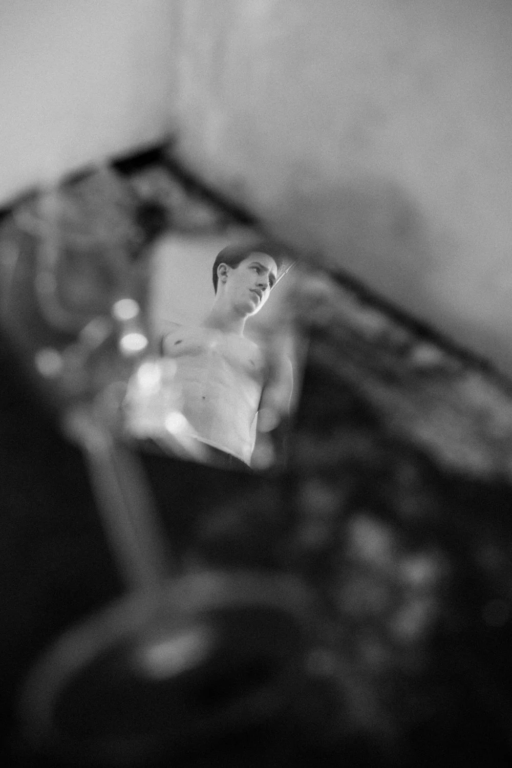 A black and white photo of a shirtless man standing in front of a mirror, with the reflection showing his face and upper body, viewed through an out-of-focus object in the foreground.