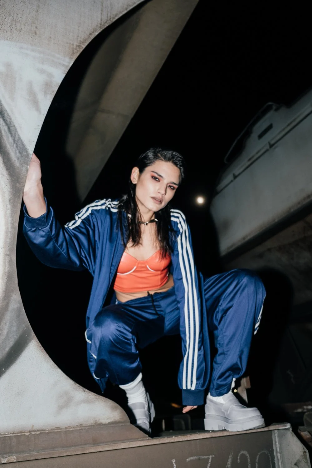 A woman in a blue athletic tracksuit and orange top squatting on a skateboard in an urban underpass at night.