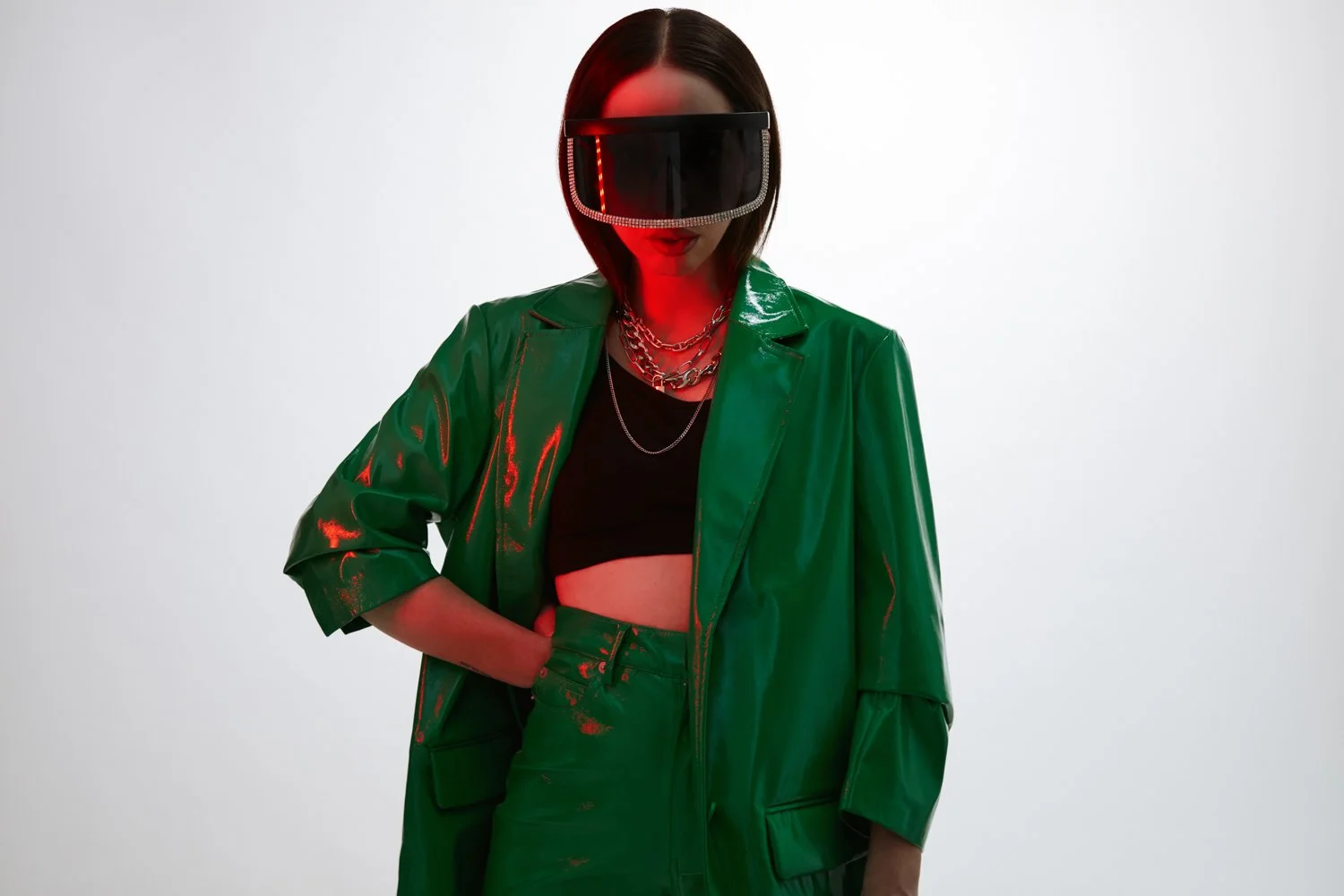Woman wearing a green leather jacket and pants, black crop top, oversized virtual reality goggles, layered necklaces, and standing with one hand in her pocket against a plain white background.