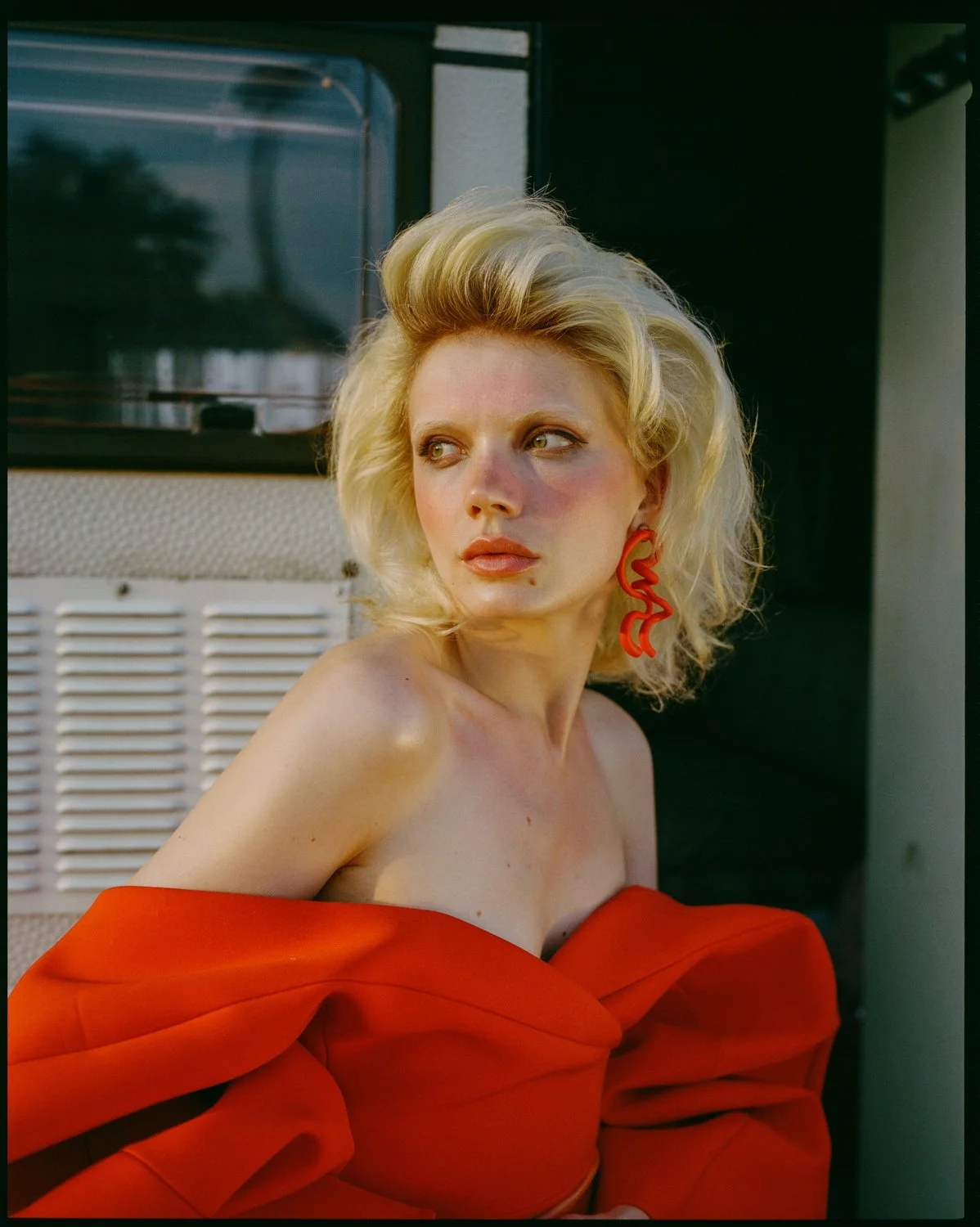 A woman with blonde hair and red lipstick wearing a red off-shoulder dress and red earrings stands outdoors near a building, looking to the side.