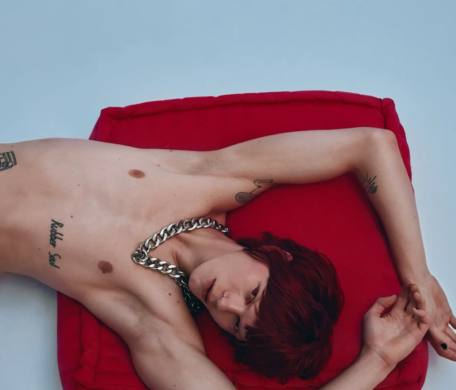 A person with red hair lying on a red cushion, shirtless, with tattoos on their arm and chest, wearing a chunky silver chain necklace, on a light background.