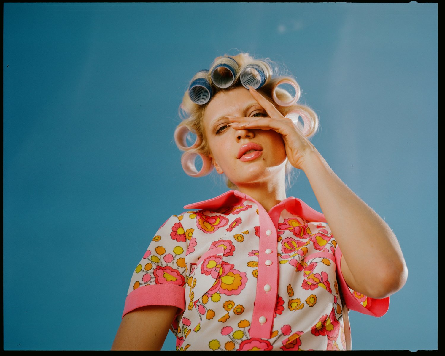 A woman with blonde hair in pink curlers, wearing a retro floral blouse, poses against a blue background, holding her hand to her face in a playful manner.