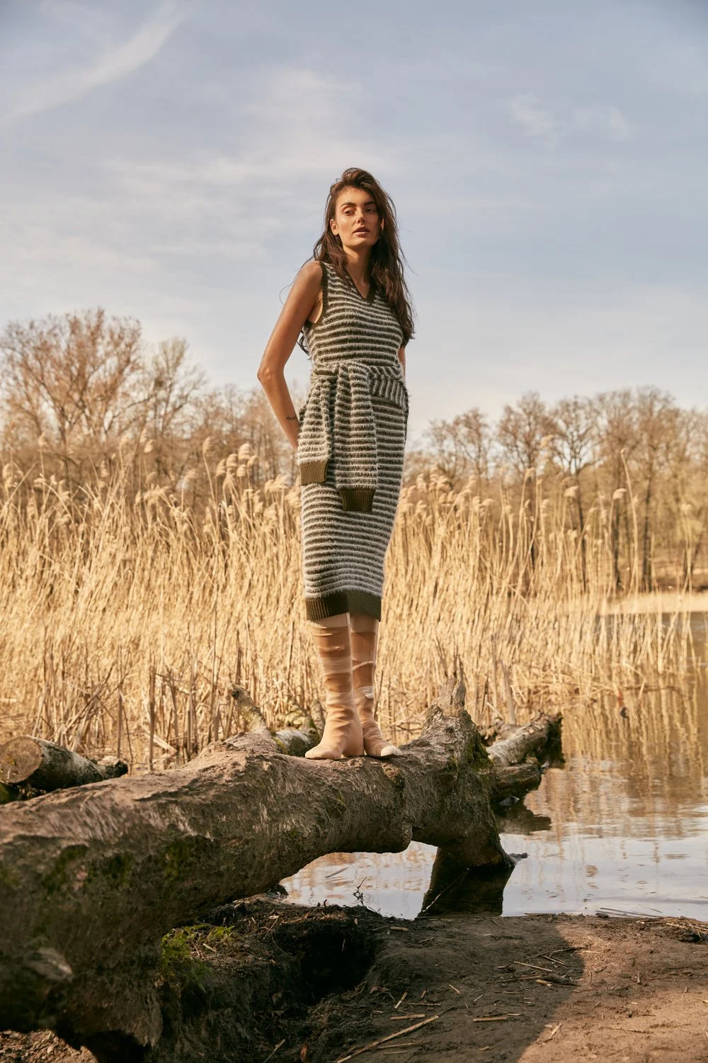 A young woman standing on a fallen tree trunk by a body of water, wearing a striped gray and black dress, matching sweater tied around her waist, and beige boots, with dry reeds and trees in the background under a blue sky.