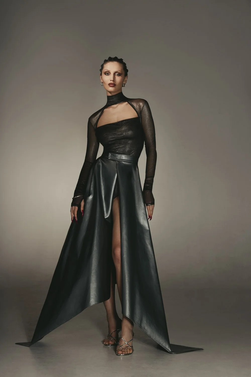 A woman in a black sheer long-sleeve top with cutout details, a high slit leather skirt, and strappy heels, standing against a neutral background.