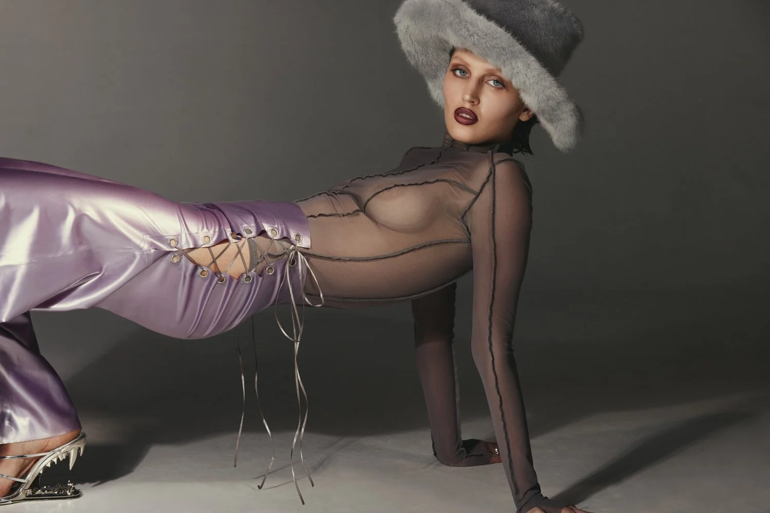 A woman wearing a large gray fur hat, sheer black top with dark piping, and shiny purple pants with lace-up sides, posing on the floor against a gray background.