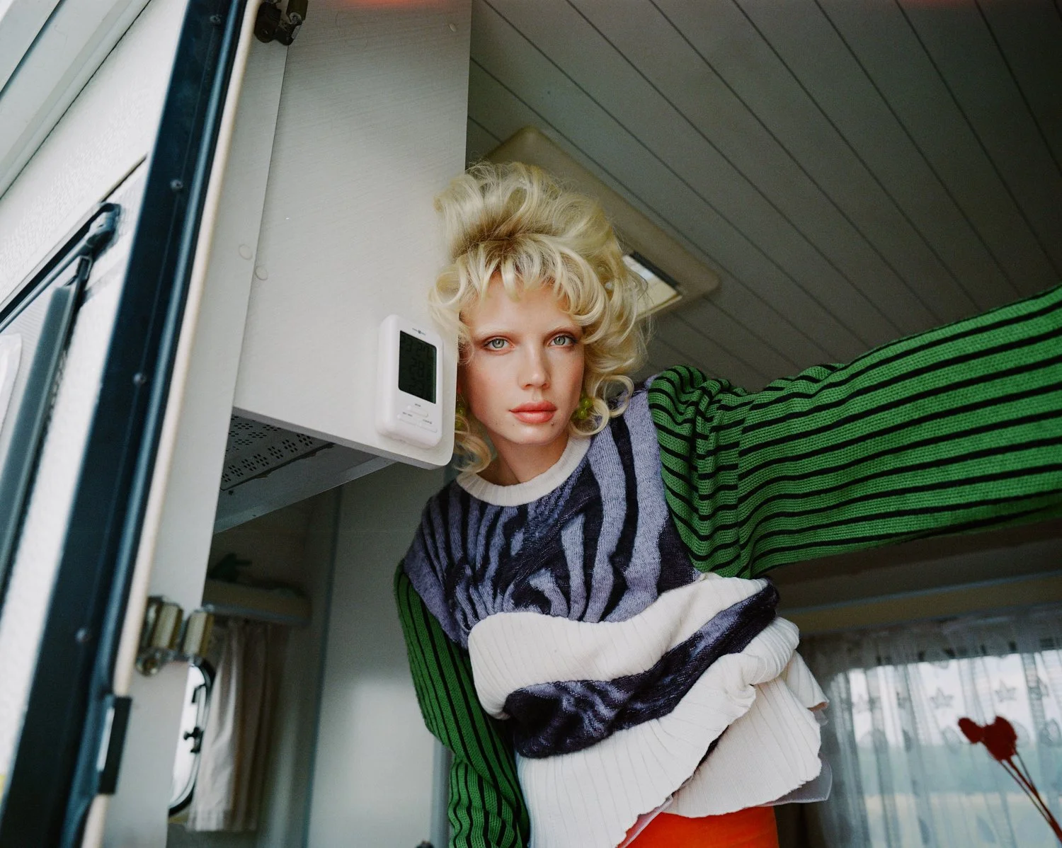 A woman with blonde curly hair and blue eyes takes a selfie inside a camper, wearing a colorful striped sweater.