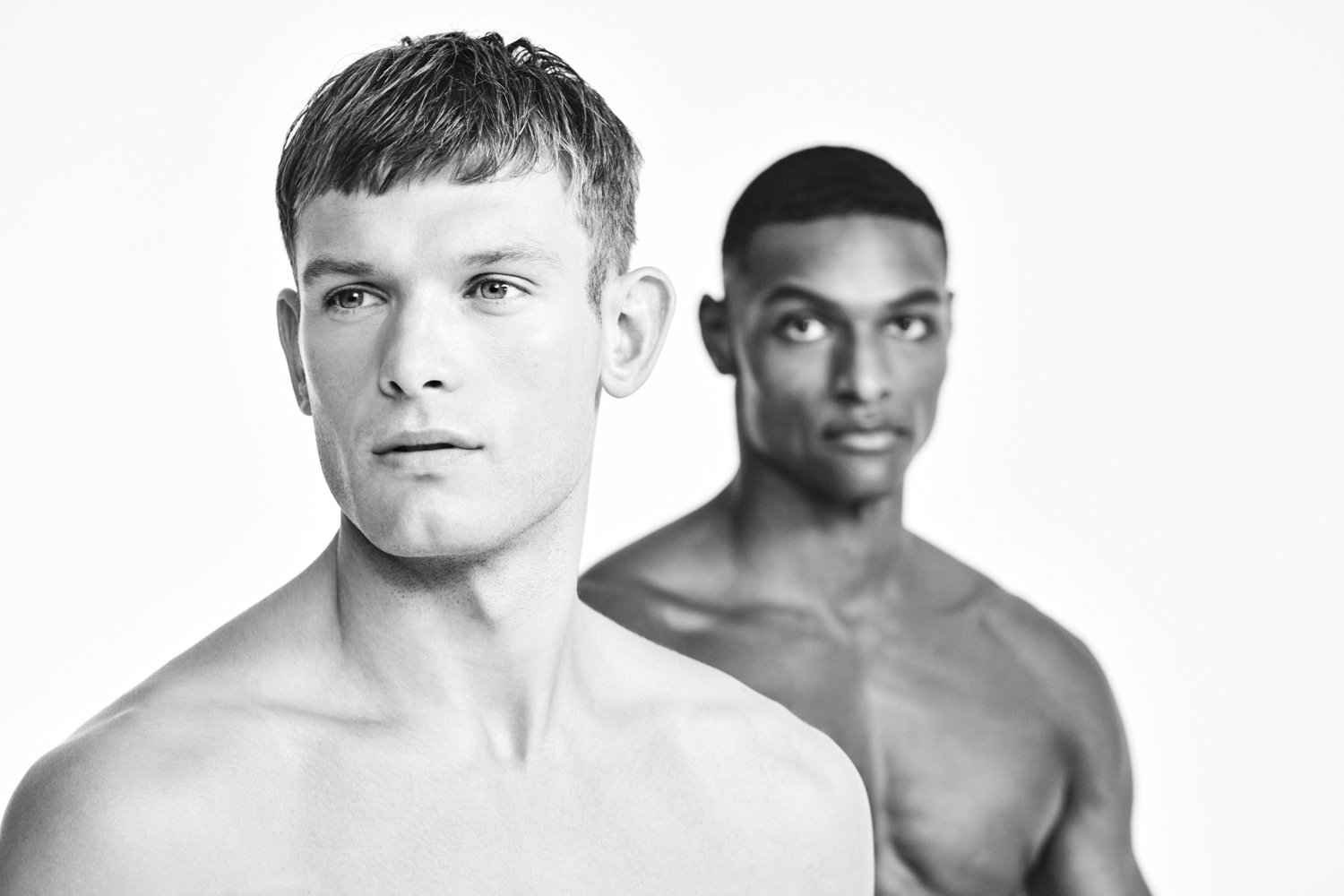 Two shirtless men, one with light skin and short hair, and the other with dark skin and short hair, pose against a plain white background.