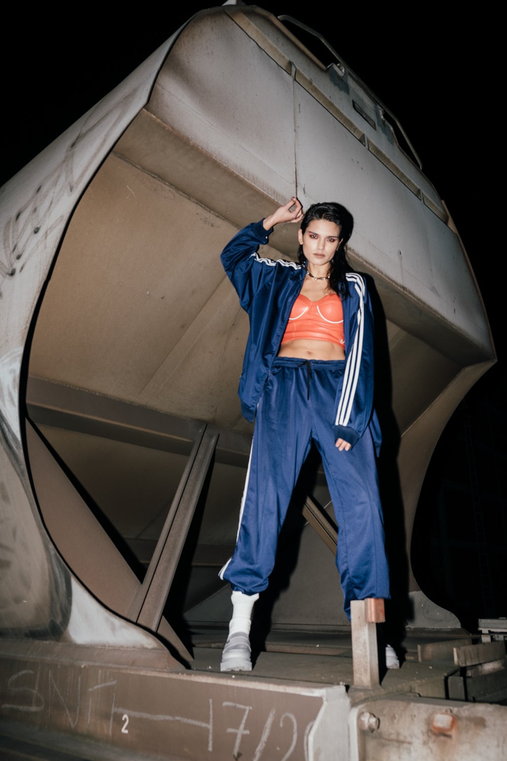 A woman standing inside a tilted, reflective, metallic structure at night, wearing a blue track suit, orange top, and white boots.