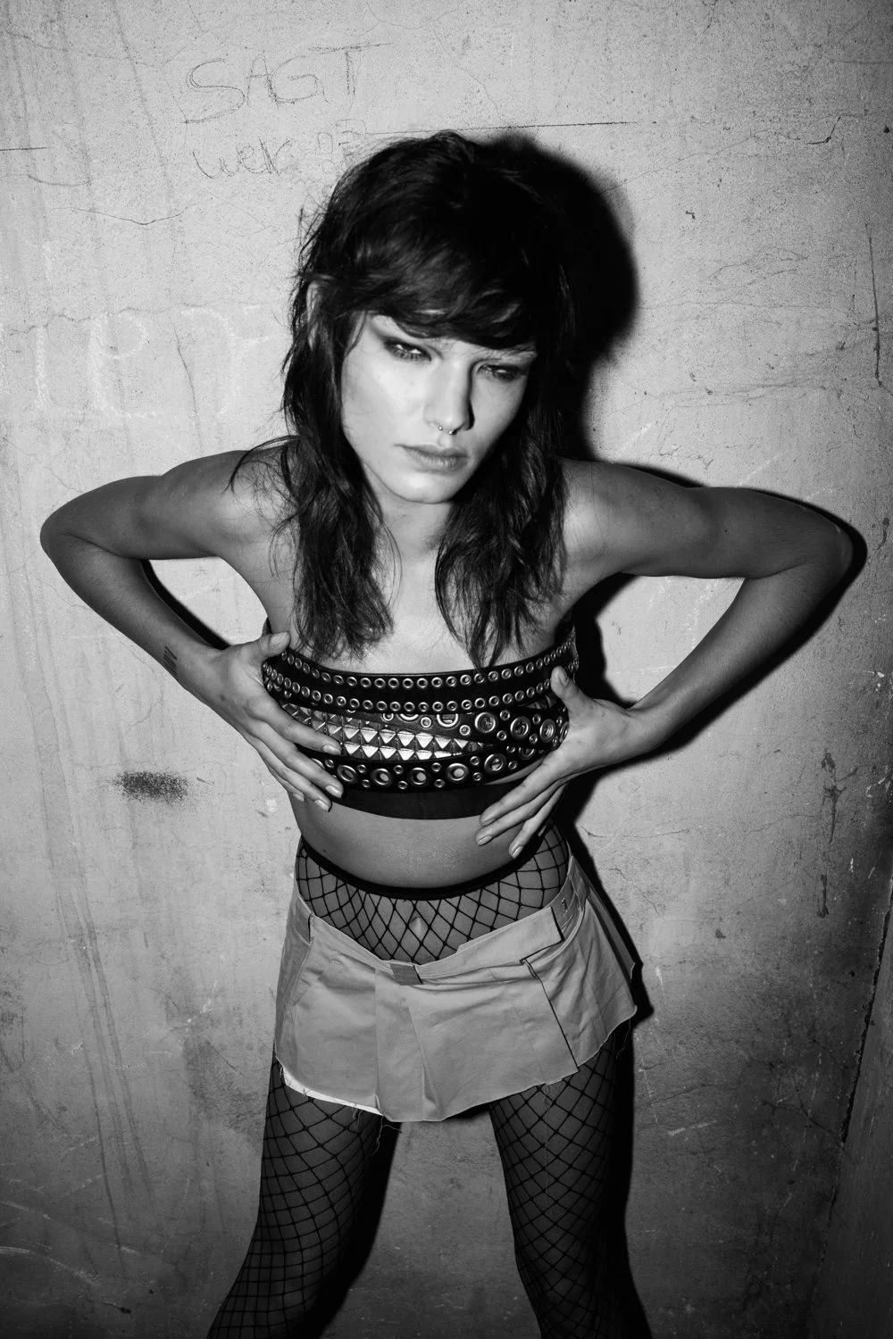A woman with dark, layered hair and a septum piercing poses against a textured wall, wearing a studded tube top, fishnet stockings, and a short skirt.
