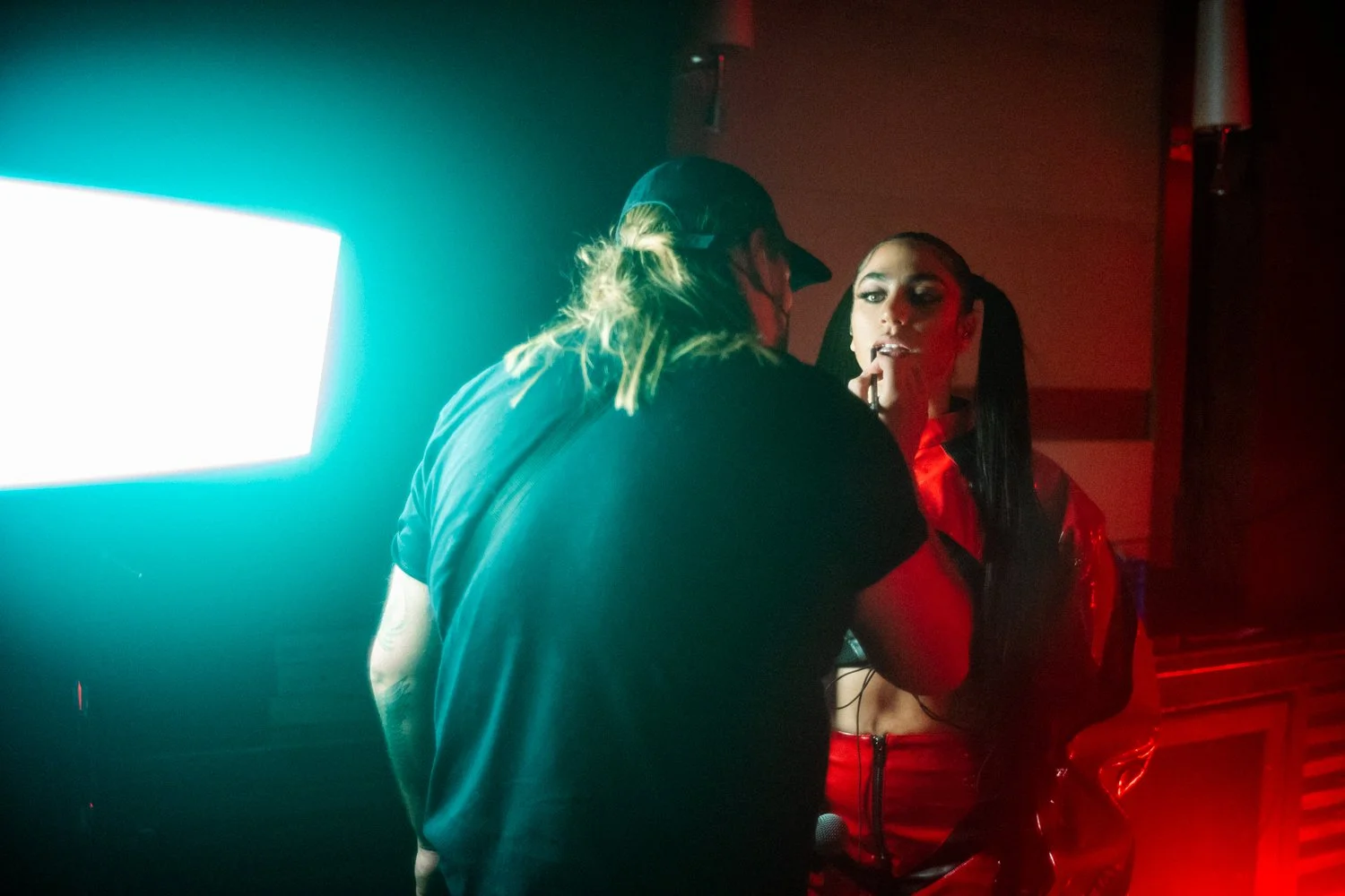 Two people, a man with long blonde hair in a backwards cap and a woman with long black hair, are in conversation in a dimly lit environment with red and blue lighting.